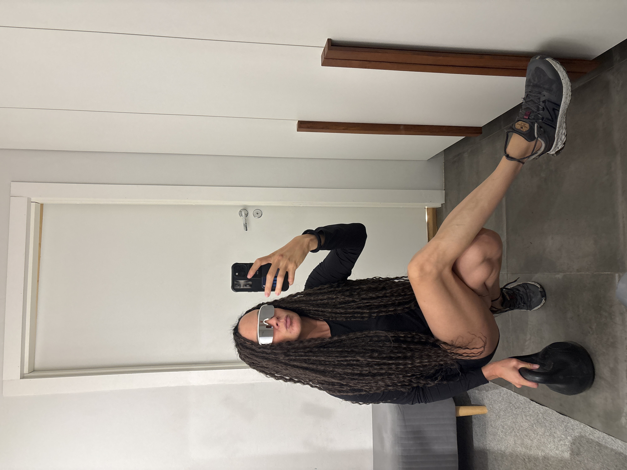A woman with long, dark dreadlocks or braids is captured in an indoor selfie, holding a black smartphone to photograph herself. She wears a long-sleeved black top and dark bottoms, complemented by mirrored rectangular sunglasses. Her attire is athletic, featuring grey and black New Balance sneakers with white soles, where the brand name "New Balance" is visible.
She is in a dynamic, squatting pose: her right leg is bent, and her left leg is extended upwards, with her shod foot resting against a white wall or door frame. Her left hand rests on the floor, holding a black kettlebell. A small, grey upholstered piece of furniture is partially visible in the lower part of the frame.
The setting appears to be a modern interior with light-colored walls, possibly white or light grey, and a dark grey concrete or tiled floor. In the background, there's a white door with a silver handle and white trim, as well as decorative horizontal wooden slats or shelves on the wall. The lighting is consistent with an indoor environment.
The image portrays a moment of a person posing with fitness equipment, likely as part of a workout routine or to showcase her athletic build and style. This scene takes place in Chaikovsky, Russia.