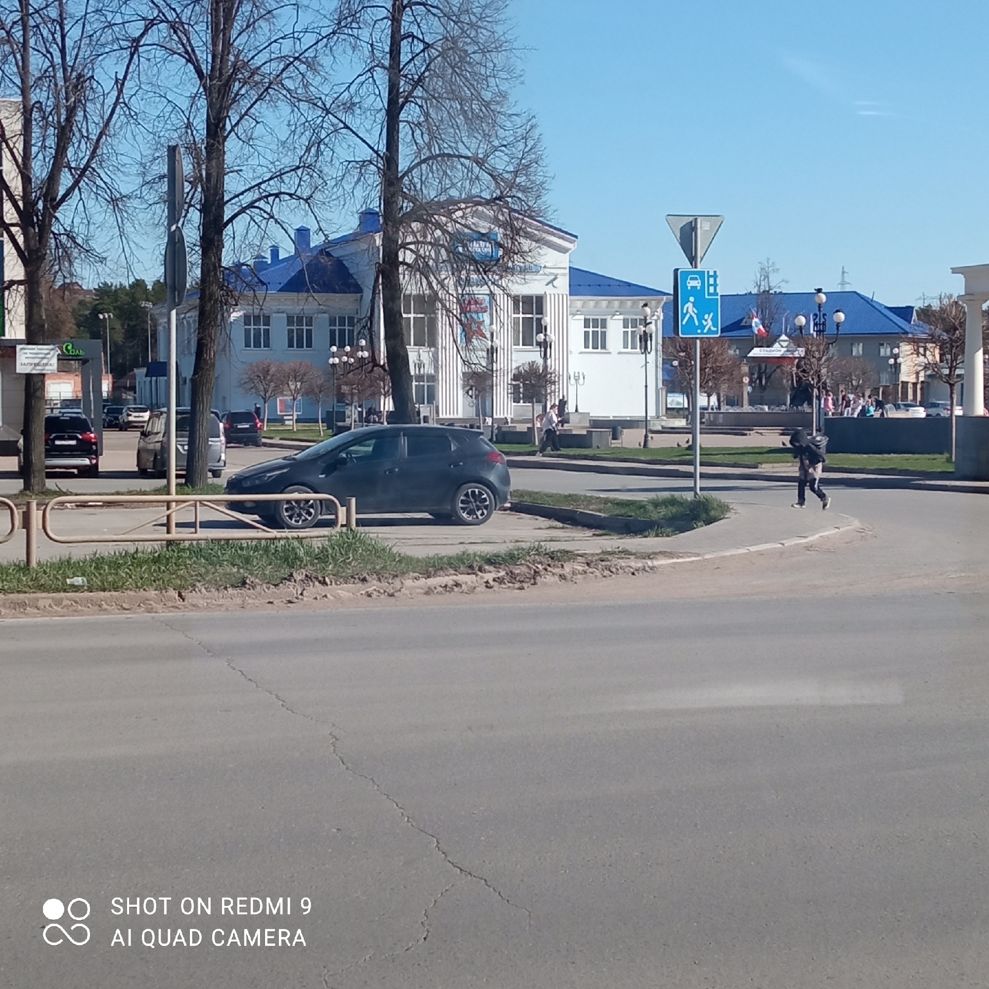 The image captures a street scene in Chaikovsky, Russia, on a sunny day. A dark hatchback car is parked beside a sidewalk with a metal barrier. A person is walking across the street at a crosswalk, indicated by a blue sign with white figures of a person and a child. In the background, a white building with a blue roof stands prominently, possibly a public building or institution given its architectural style. Several bare trees line the street and area around the building. Further in the distance, other cars are parked and pedestrians are visible. The foreground is dominated by a gray asphalt road with cracks. The sky is clear and blue. The lower part of the image contains a watermark: “SHOT ON REDMI 9 AI QUAD CAMERA."