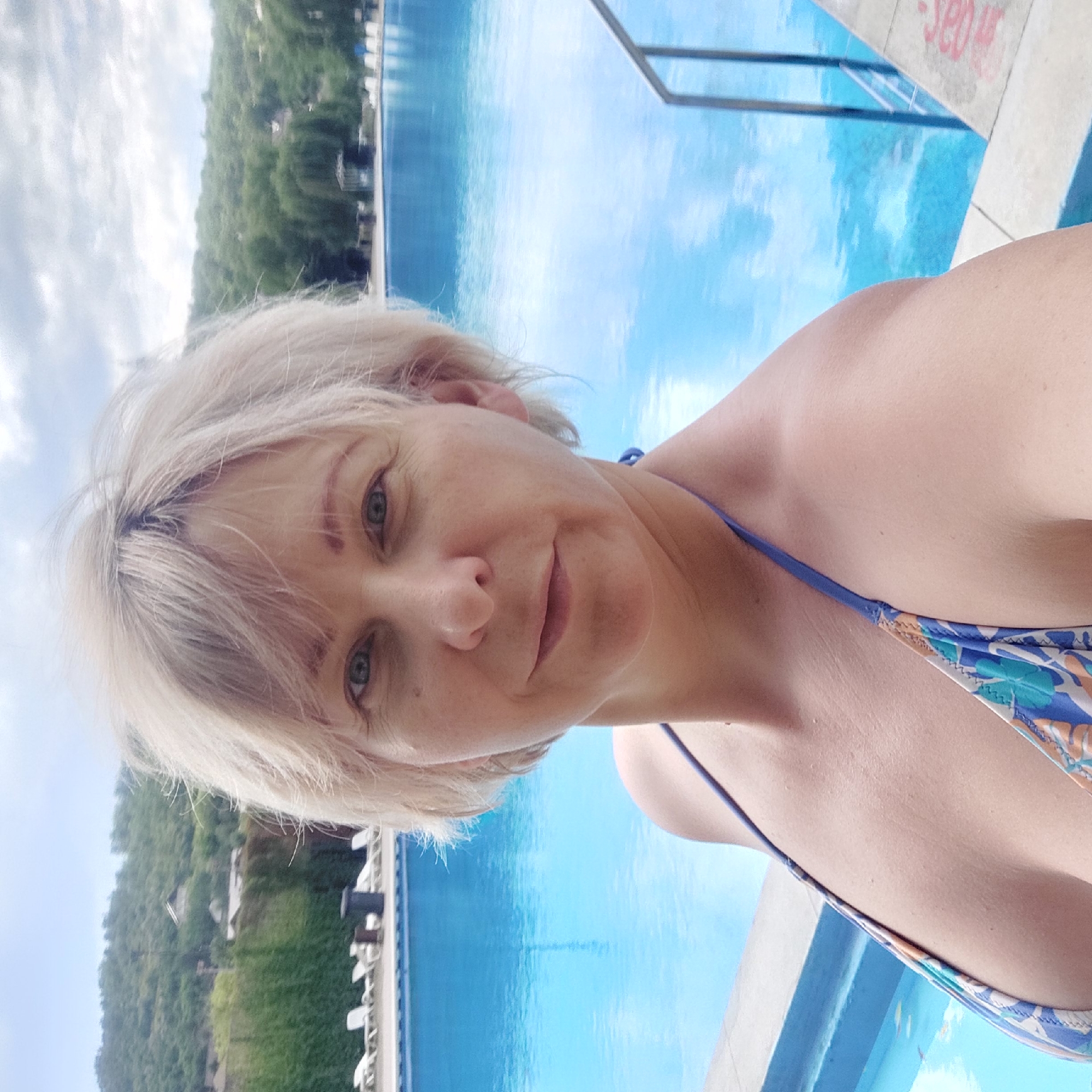 A woman with short, blonde hair and blue eyes takes a selfie in front of a swimming pool. She is wearing a patterned bikini. The pool is large and blue, with lounge chairs visible in the background. Beyond the pool, there is a green, treed hillside with some buildings on it. The sky is cloudy. The location is a swimming pool in Iași, Romania.