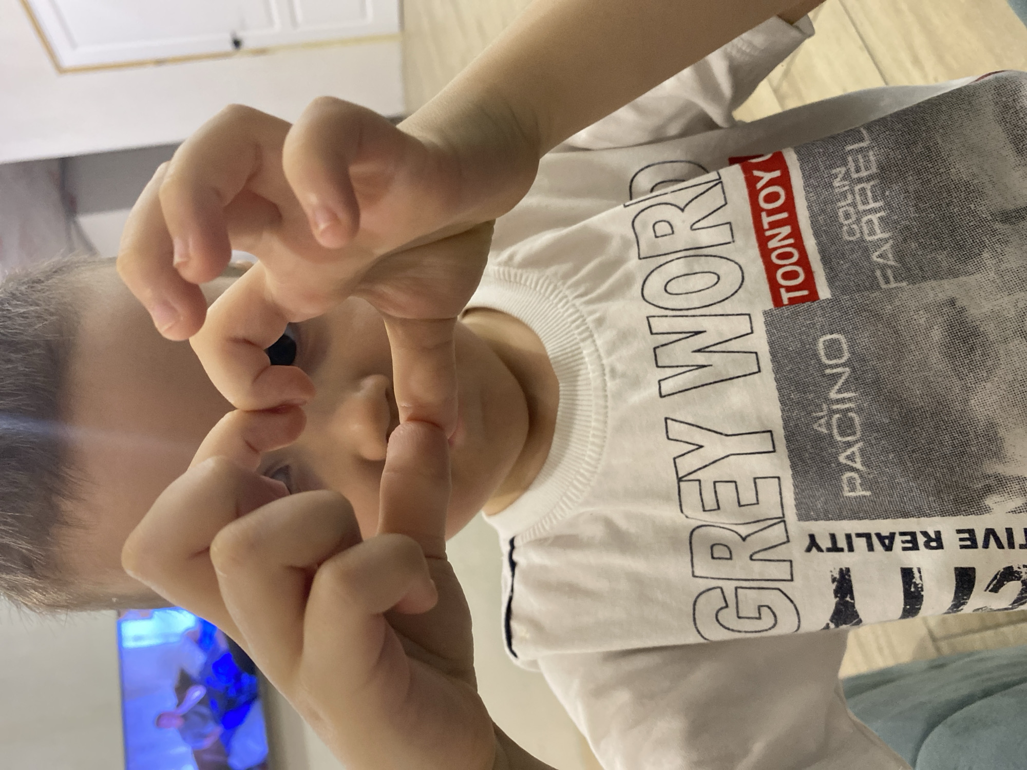 A child with fair skin and short, light-colored hair is positioned close to the camera, forming a heart shape with their hands around their eyes. They are wearing a white t-shirt printed with the words "GREY WORD TOONTOY COLIN FARREL" and "AL PACINO," and a graphic design. In the background, a television screen displays an image with blue and white hues. The setting appears to be indoors, with a glimpse of light-colored flooring and white cabinetry near the top of the frame. There's no information given about the location in the city of Chaikovsky, Russia.