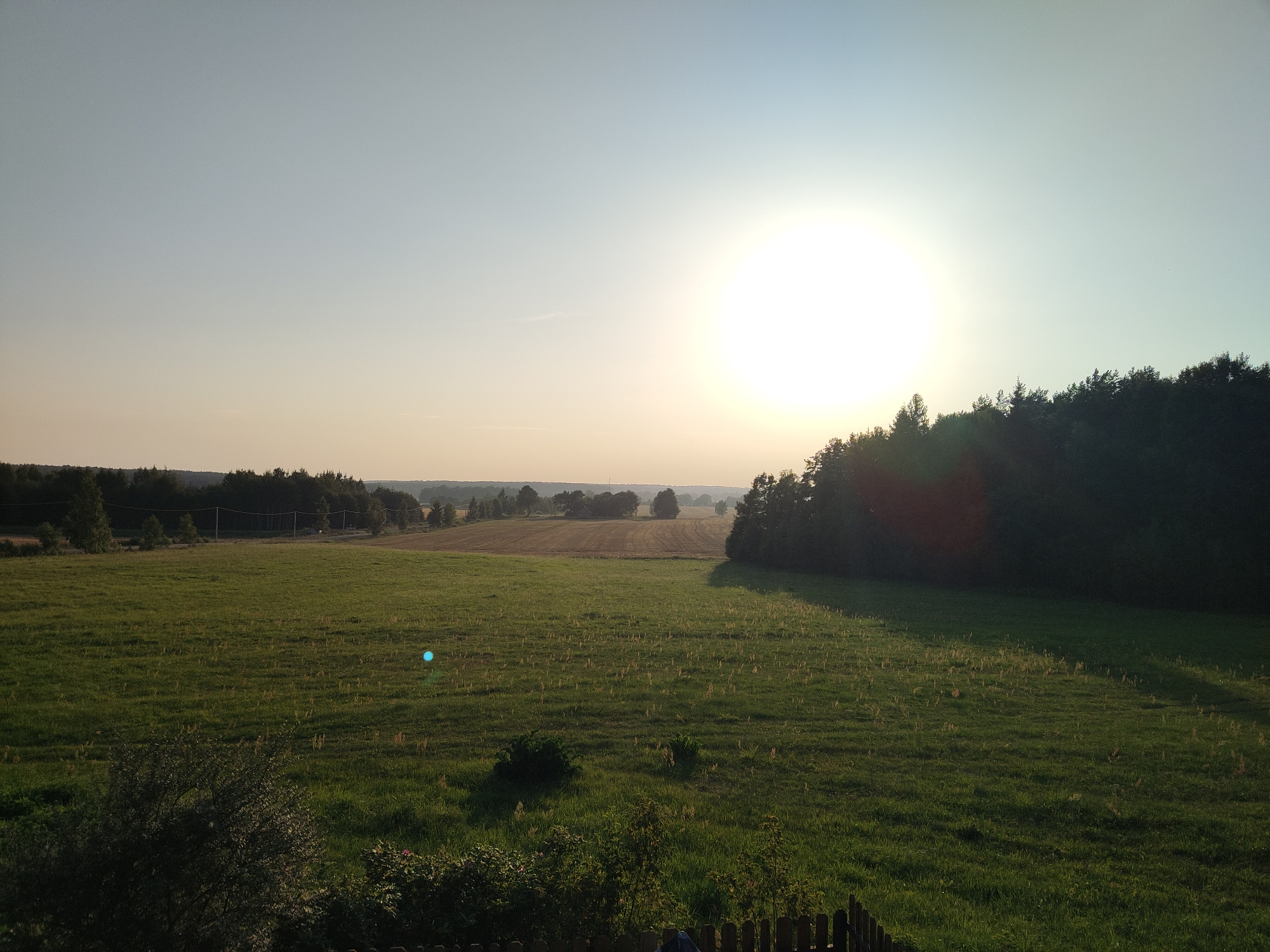 The image depicts a sunlit landscape with a vast expanse of green fields and a distant tree line under a clear sky. The sun, a bright disc, is positioned high in the sky, casting a warm glow. A small, circular blue lens flare is visible in the lower left quadrant of the field. In the foreground, there is a dark wooden fence, partially obscured by foliage. The environment appears to be rural, consistent with the location of Norviliškės, Lithuania, known for its pastoral scenery. There are no people or discernible actions captured in the image.