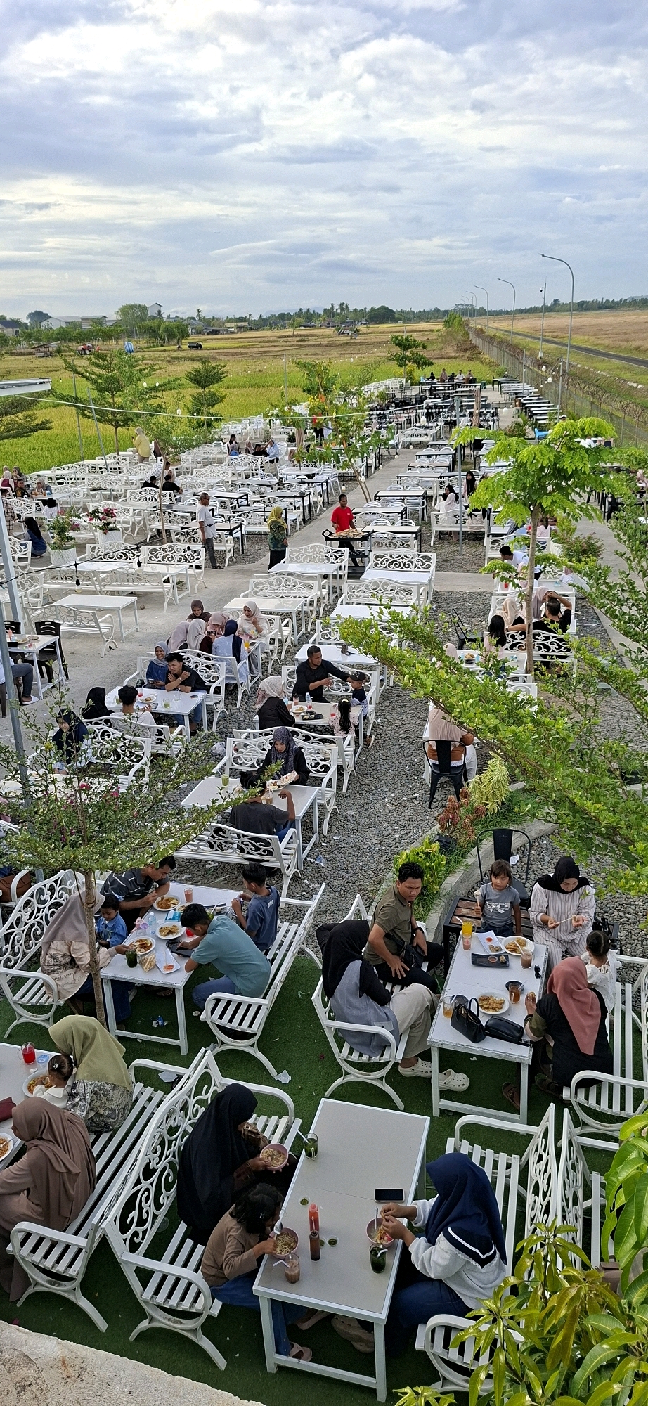 The image shows an outdoor dining area at a restaurant located in Buengpageu, Indonesia. The restaurant has many white tables and chairs set up on a gravel and green artificial turf surface. Several groups of people are seated at the tables, eating and socializing. The background reveals a rural landscape with fields and trees. The sky is cloudy. The restaurant appears to be a popular spot with a large number of patrons.