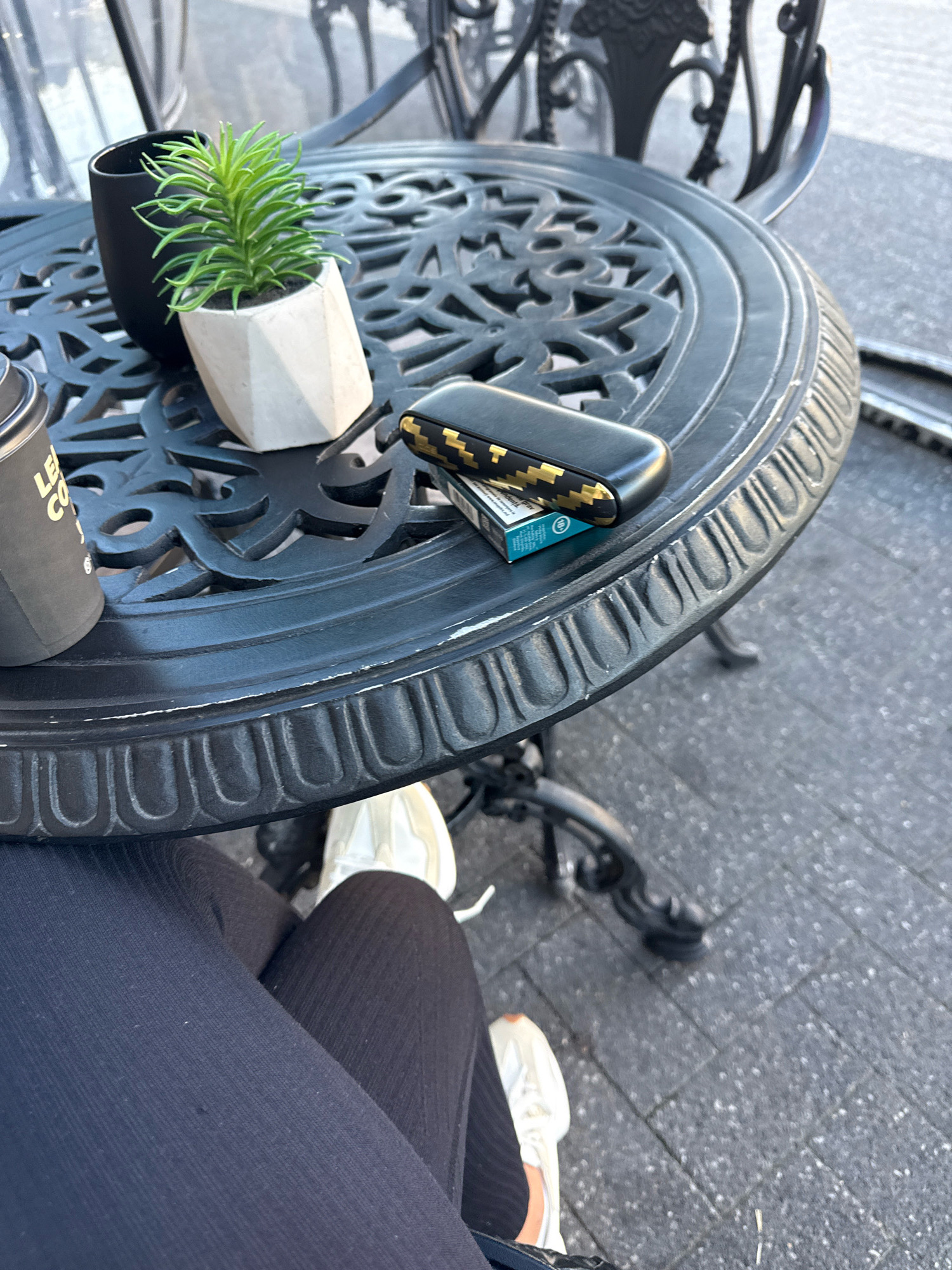 A person's legs are visible, wearing black ribbed leggings and white sneakers, sitting at a black metal table on an outdoor paved surface. On the table are a black mug, a small potted succulent plant in a geometric white planter, a pack of cigarettes, and a black and gold patterned electronic cigarette device. The table appears to be part of a cafe or restaurant setting. The lighting suggests it is daytime. The background shows more black metal table and chair structures, with a blurred outdoor environment. There is no direct indication of the location being Chișinău, Moldova, but the style of the furniture and paving is consistent with urban outdoor cafe settings.