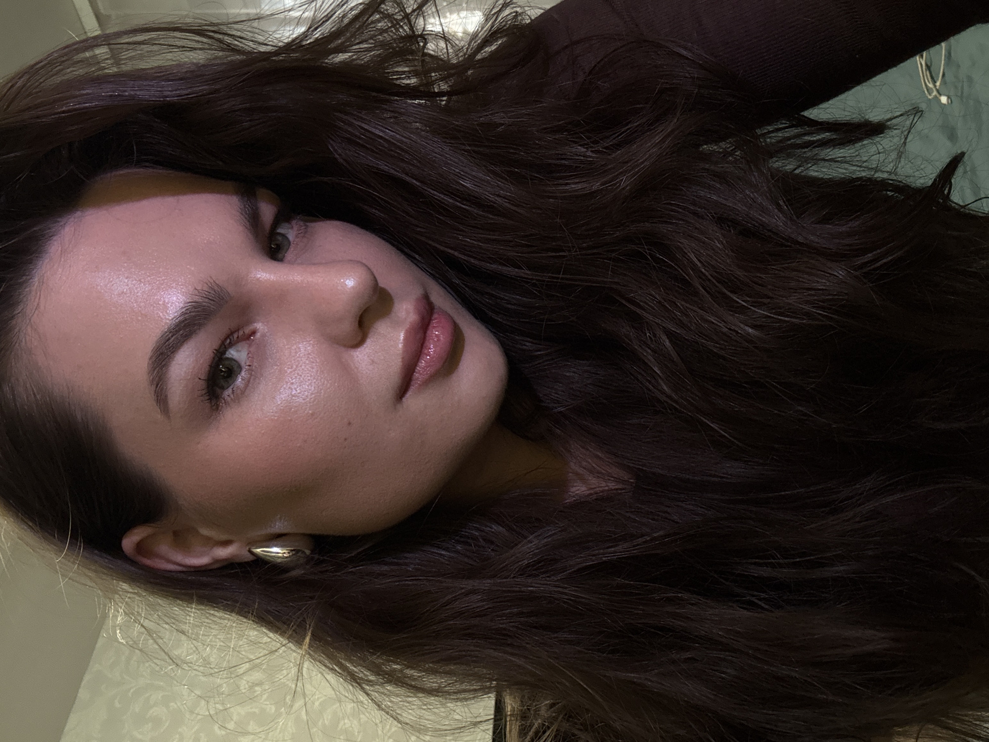 The media file is a close-up, portrait-oriented photograph of a young woman with long, dark brown, wavy hair. Her face is tilted, and she is looking towards the viewer with a neutral expression. She is wearing makeup, including lipstick and what appears to be highlighter on her cheeks and nose, giving her skin a dewy appearance. She has dark eyebrows and her eyes are open. She is wearing a dark brown top. A large, silver, tear-drop-shaped earring is visible in her left ear. The background is blurred, but suggests an indoor setting with patterned wallpaper on the left and a dark green surface on the right, possibly a wall or furniture. The lighting appears to be artificial, casting soft shadows and highlighting the texture of her hair and skin. There is no visible text in the image. The image does not provide specific clues about the time of day, weather, or a precise location within Chaikovsky, Russia, beyond being an indoor setting.