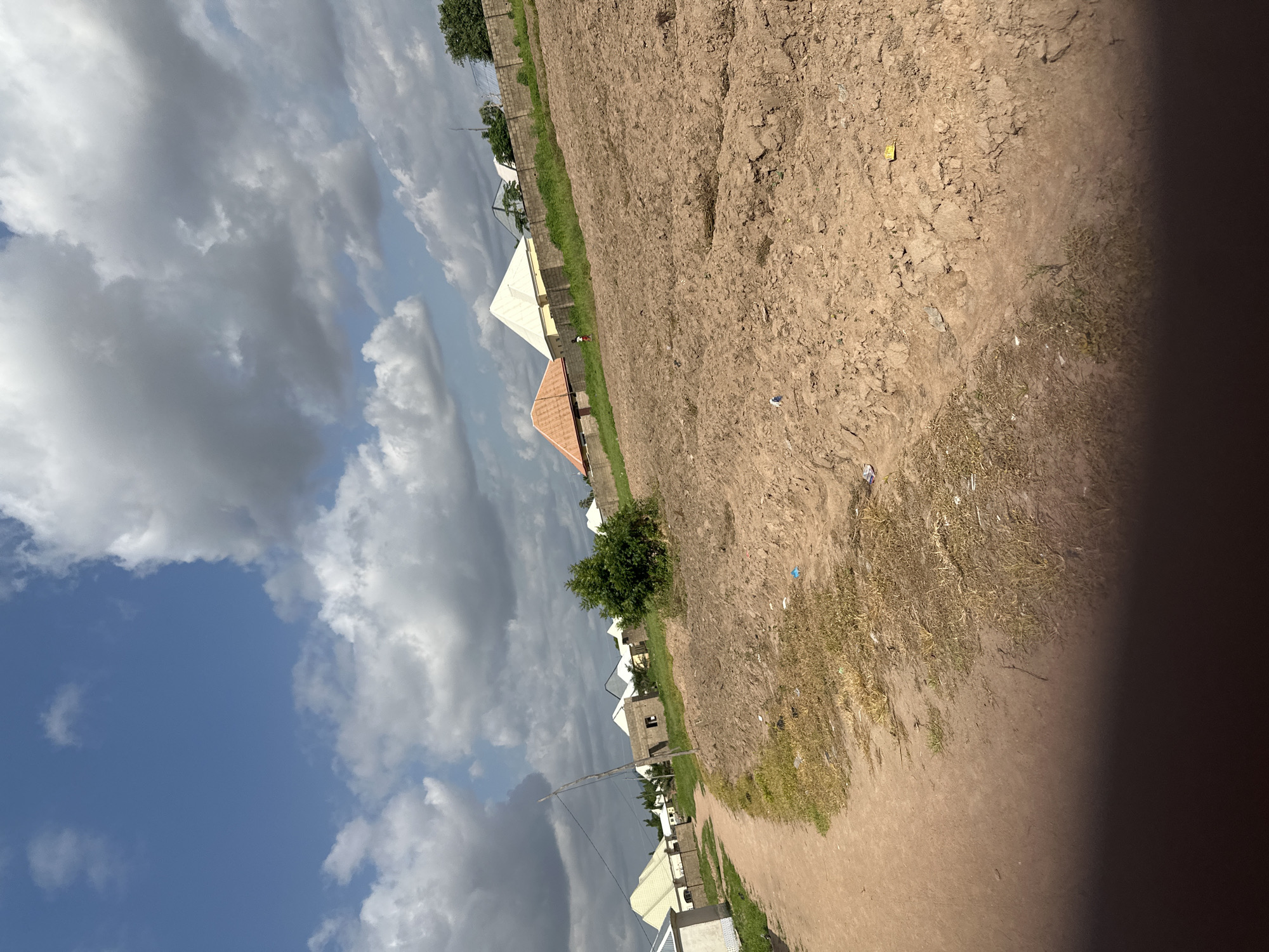 This image depicts a rural or semi-urban scene in Jalingo, Nigeria, under a partly cloudy sky. The perspective is from a slightly elevated angle, looking out over a landscape that combines natural elements with signs of human habitation and development.
The primary subjects are a series of buildings, many of which appear to be under construction or newly built. These structures feature distinctive triangular roofs, some white and others with an orange, tiled appearance. The construction suggests ongoing development in the area. In the distance, a low brick wall runs along the edge of what appears to be a cultivated field or open plot of land. The field itself is characterized by dry, brown soil, with patches of sparse, dry grass.
A small number of people are visible, appearing as small figures in the distance, suggesting the scale of the buildings and the open space. One small figure is standing near a building.
The weather appears to be dynamic, with large, dramatic clouds, some dark and heavy, indicating potential for rain, interspersed with patches of bright blue sky. The lighting suggests it is daytime, likely late morning or afternoon, given the shadows and the brightness of the sky.
Notable details include the presence of dry vegetation, indicating a possibly arid or dry season climate, and the debris scattered on the ground, such as small pieces of paper or plastic, which are common in many outdoor environments. The overall impression is one of a developing community in a somewhat dry landscape. There is no visible text in the image.