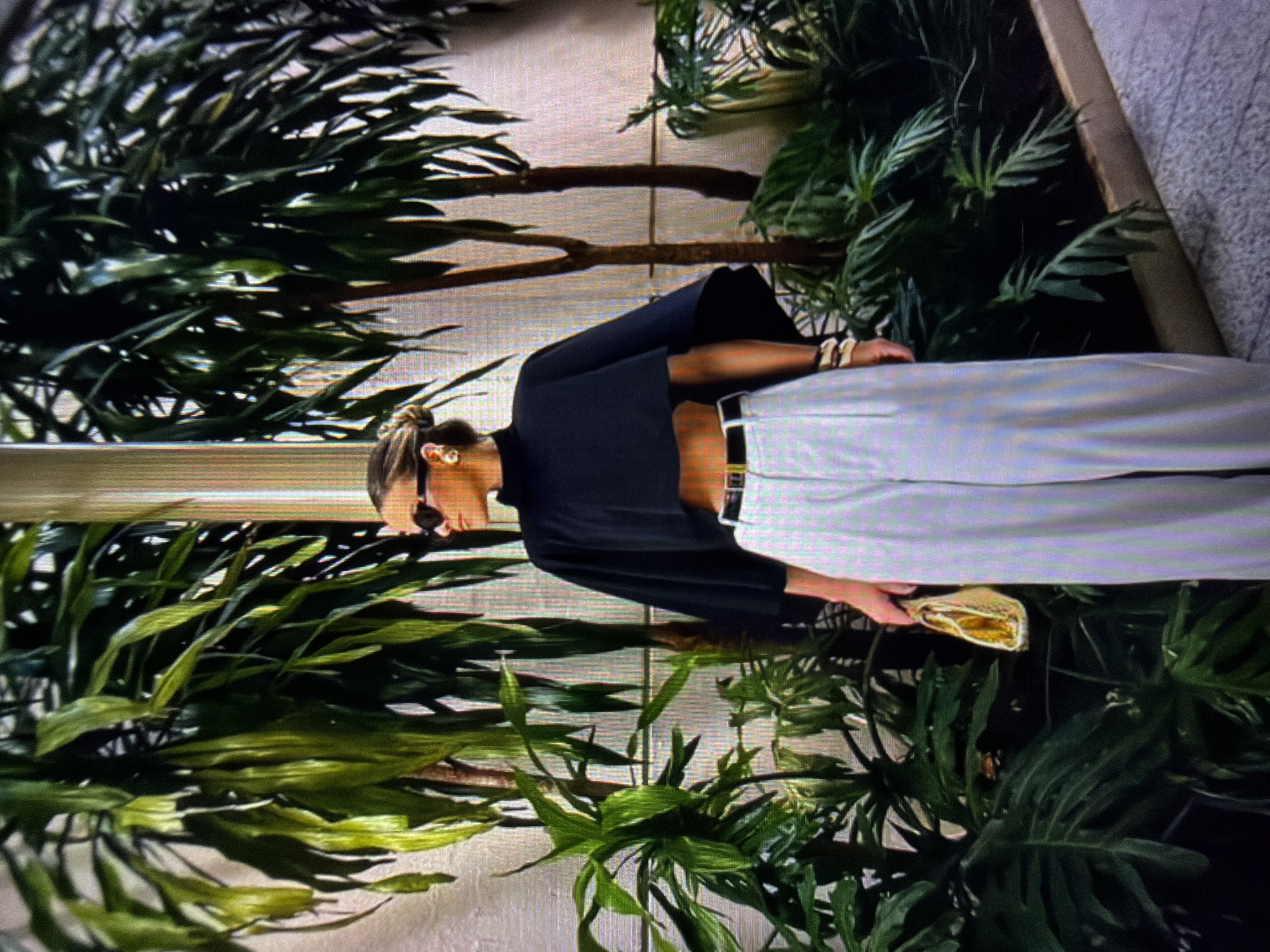 The image shows a woman standing outdoors, surrounded by lush green plants. She is wearing a black, cropped, high-neck top and wide-leg, light-colored pants. She has sunglasses on and is holding a small, textured yellow clutch bag in her left hand. Several bracelets adorn her right wrist. The background consists of tall potted plants with long, slender leaves, and some plants with larger, broad leaves. A light-colored wall is visible behind the plants. There is a narrow, raised planter edge in the foreground, and a textured ground surface, possibly a walkway or patio, is visible at the bottom. The scene appears to be a well-maintained outdoor space, likely a botanical garden, courtyard, or a similar urban green space within a city. Given the context of the prompt mentioning Beșalma, Moldova, it is plausible this is an urban outdoor setting in that city, possibly a commercial or public area designed with extensive foliage.