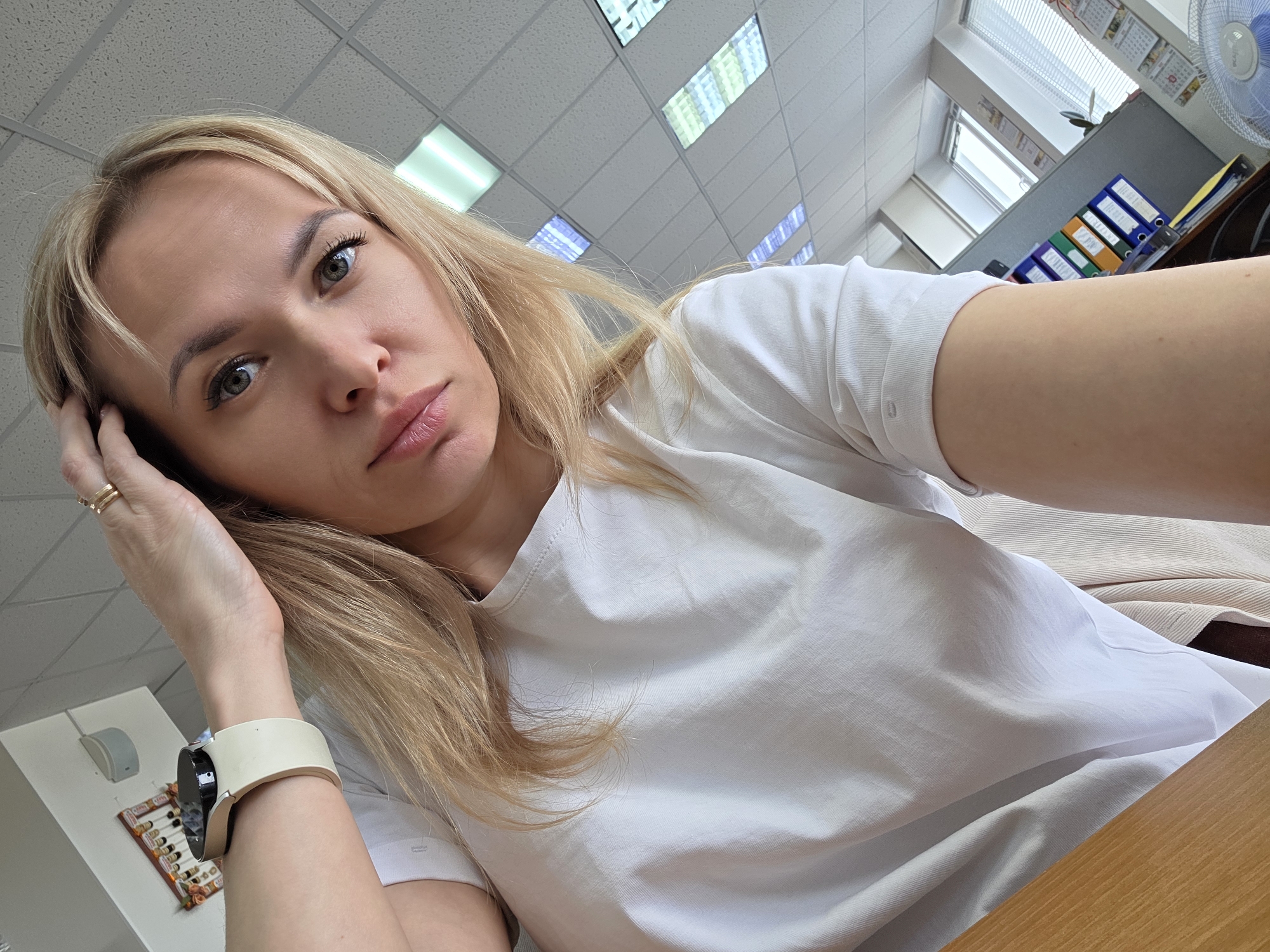 The image shows a blonde woman in a white t-shirt, likely in an office setting in the city of Chaikovsky, Russia. She is holding her head with her hand, wearing a watch on her wrist, and appears to be taking a selfie. The office includes a ceiling with fluorescent lights, windows with blinds, a desk, a fan, and binders. A calendar and other small items are visible on the wall.