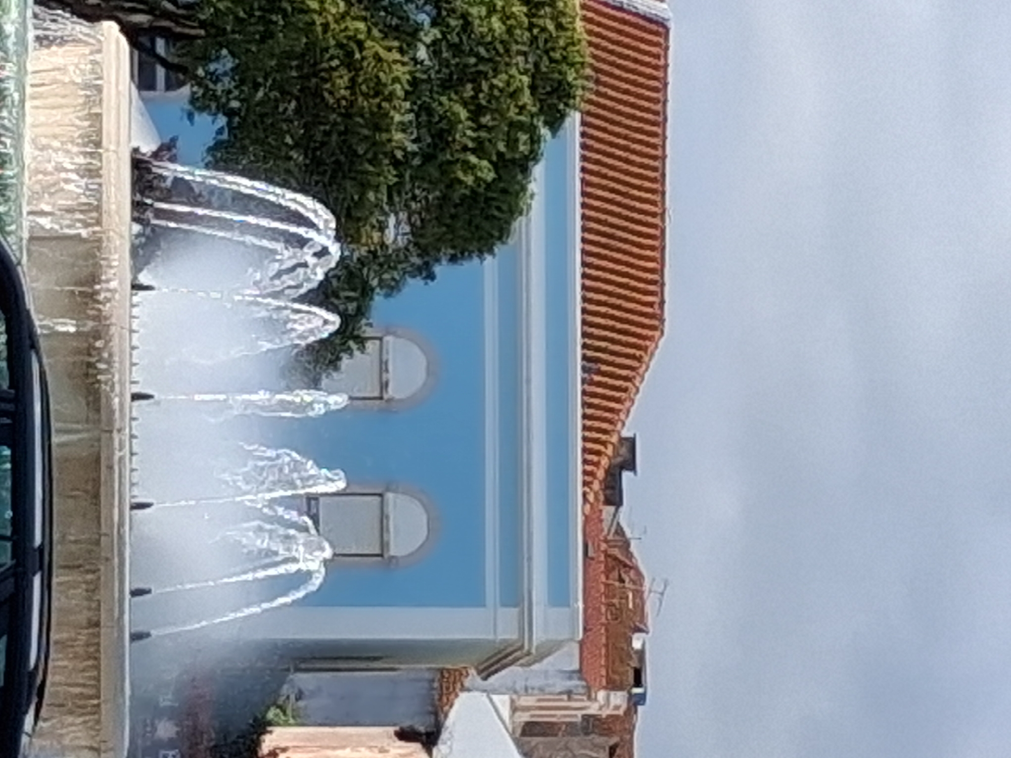 The image shows a fountain in front of a building with a blue facade and red tile roof in Figueira da Foz, Portugal. The fountain is multi-tiered and shooting water jets upwards. A tree is partially visible behind the fountain. Part of a vehicle is visible on the left edge of the image. The sky is overcast.