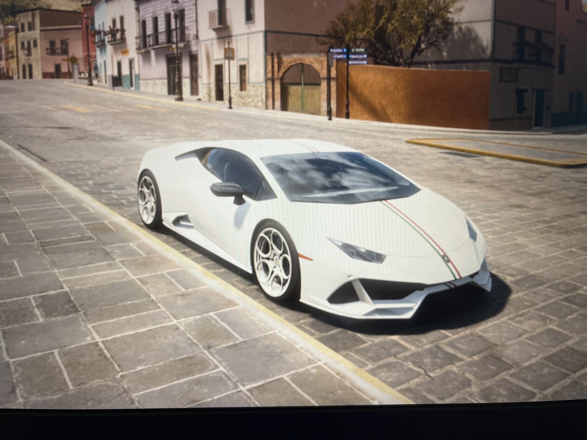 A white Lamborghini sports car is parked on a cobblestone street in front of buildings. The car has green and red racing stripes down the center of the hood. The buildings appear to be old-fashioned with balconies and arched doorways. The street is lined with a yellow curb. The scene is set in a city environment, likely on a sunny day. The location is not Varna, Bulgaria, as there are no identifying landmarks or features that associate this scene with that city.