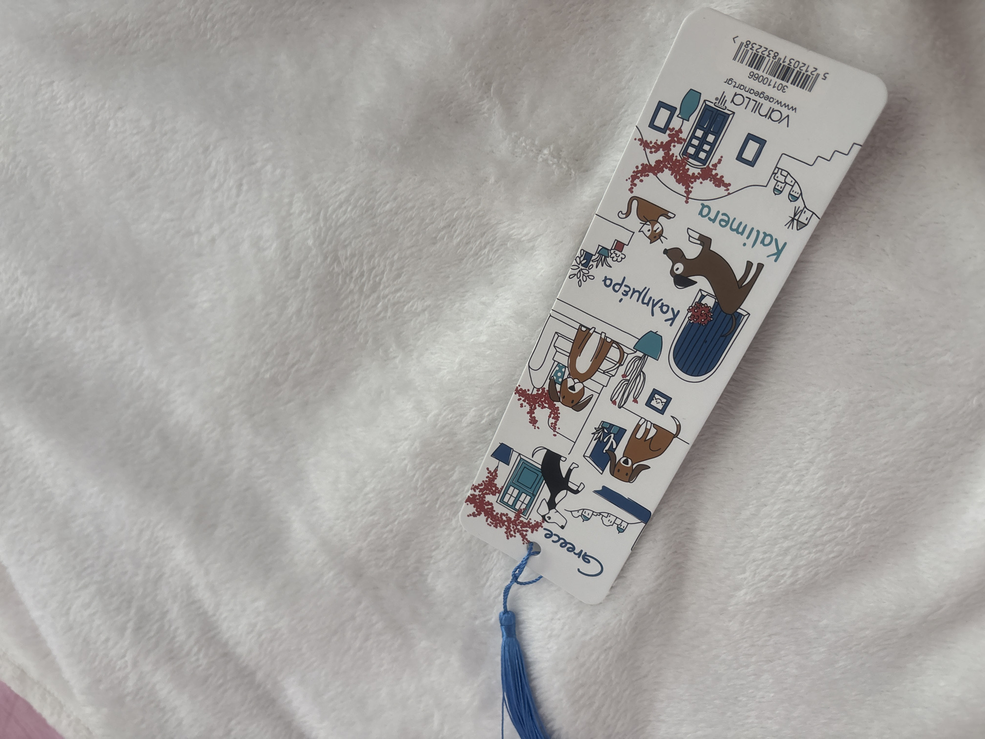 A bookmark with a blue tassel lies on a white, fluffy blanket. The bookmark features a whimsical illustration of Greek village scenes, including dogs, cats, and traditional houses. The word "Greece" is written on the left side, and "Kalimera" (meaning "good morning" in Greek) is written in the center. On the right side, there is a barcode and the text "Vanilla www.aegeansart.gr 30110066 5 212031 832238 >". The bookmark is positioned diagonally across the lower portion of the frame. The background is entirely composed of the soft, white blanket, which is textured and appears to be a plush fabric. There is no indication of Strelcha, Bulgaria in the image.