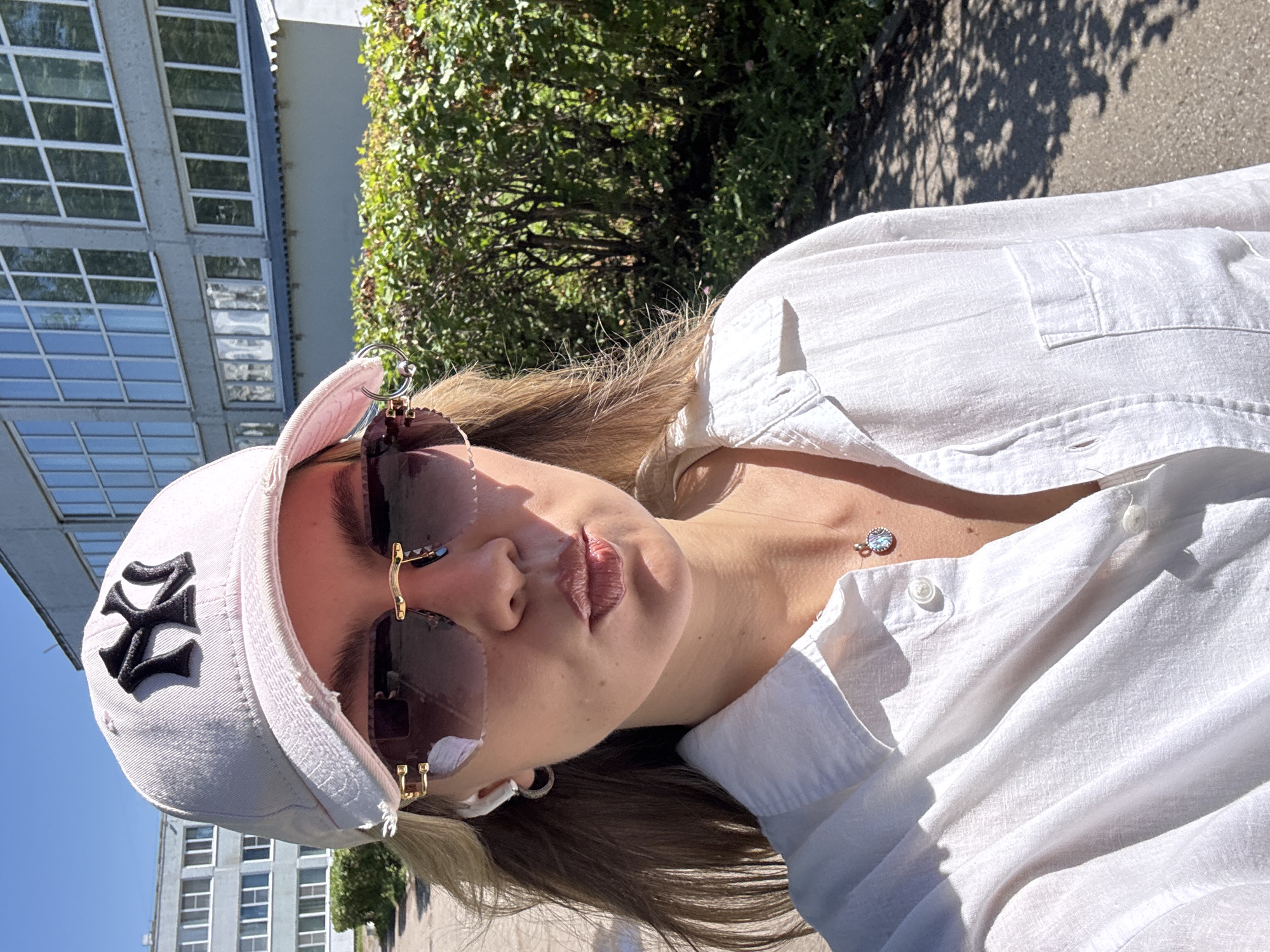A young woman, wearing a white New York Yankees baseball cap, oversized sunglasses with gold accents, and a white linen shirt, poses for a selfie outdoors. She has on small hoop earrings and a necklace with a blue pendant. The background consists of a building with large windows and lush green bushes. The scene is lit by bright sunlight, casting shadows on the paved walkway. The woman's lips are slightly puckered, and her hair is flowing.