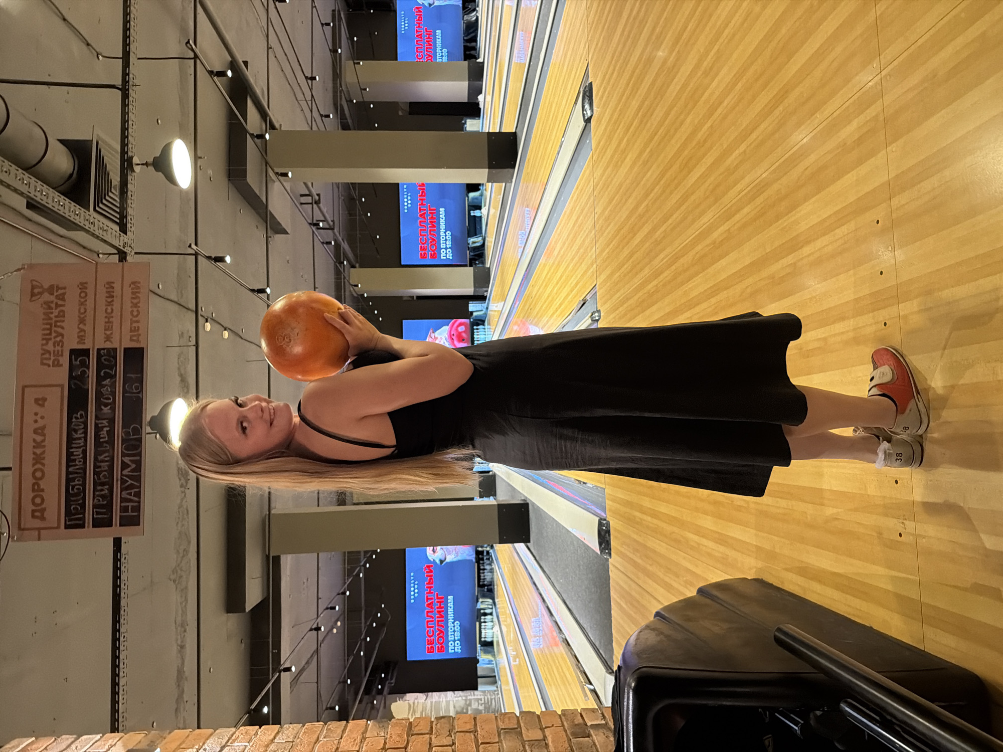 The image shows a woman holding an orange bowling ball, standing on a wooden bowling lane. She is wearing a black dress and red and white bowling shoes. In the background, there are multiple bowling lanes and screens displaying advertisements. Above the bowling lanes, a scoreboard shows rankings. The overall environment suggests an indoor bowling alley. The image itself does not contain any explicit information to determine the city or country.