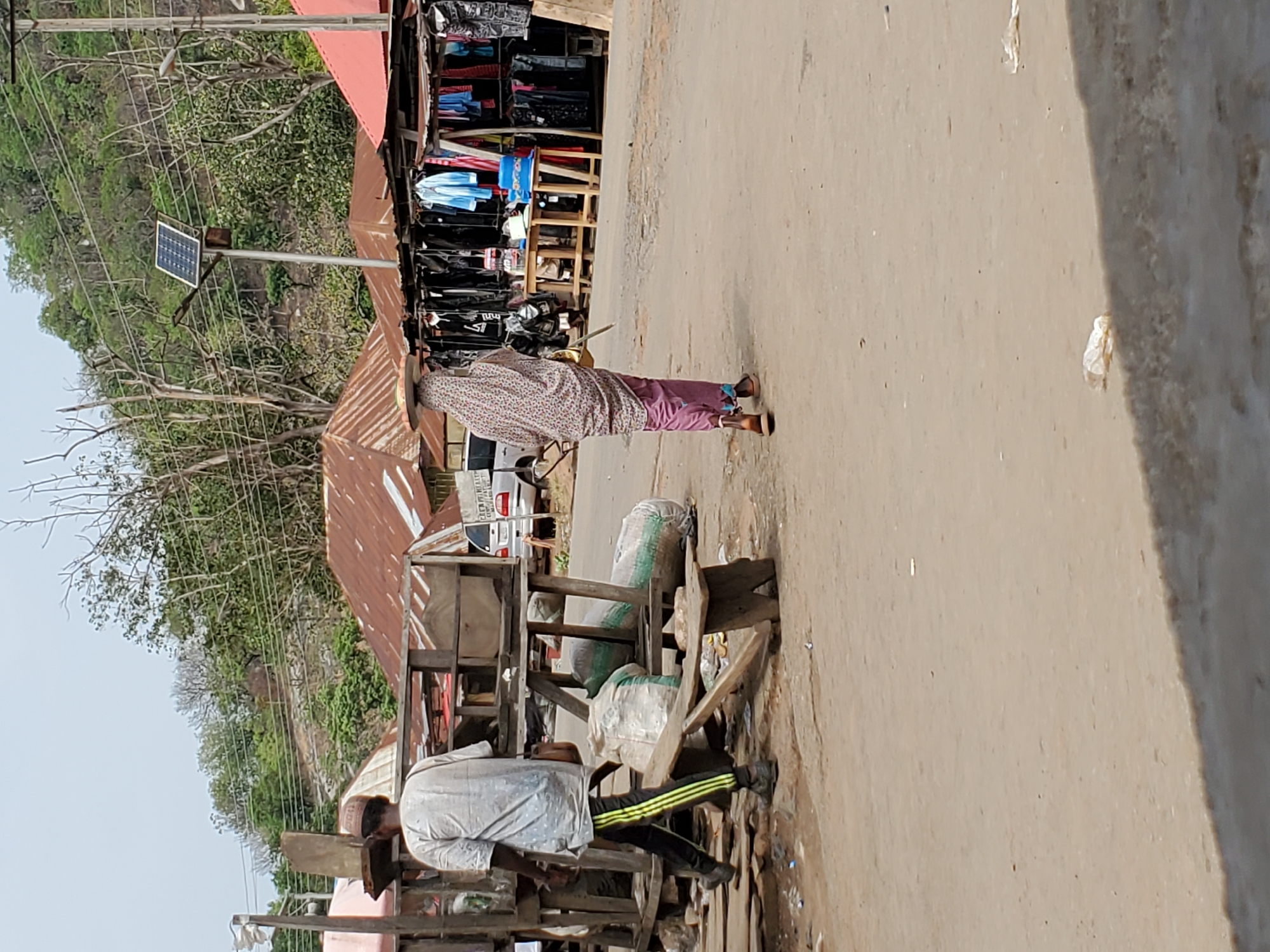 The image shows a street scene in Mararaba, Nigeria. Two individuals are visible near makeshift wooden structures that appear to be market stalls. One person, dressed in a patterned shirt and pink pants, is standing on the road. The second person, wearing a light-colored shirt and pants with stripes, is near a stall laden with sacks. A solar panel is visible on a pole nearby. The road surface is light brown. In the background, there are trees and buildings with rusted roofs. Clothes are hung on racks near the stalls. A car is parked in the background.
