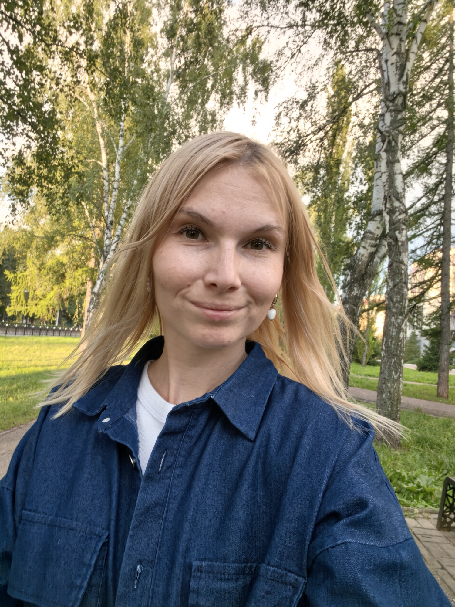 A blonde woman with fair skin and brown eyes is captured in a selfie outdoors. She is wearing a dark blue denim jacket over a white t-shirt and a single white earring with a heart shape. Her hair is long and light blonde, gently blowing in the wind. The background suggests a park or green space, with birch trees and sunlight filtering through the leaves, indicating it is likely daytime. The scene appears to be in a city, possibly Sterlitamak, Russia, given the context.