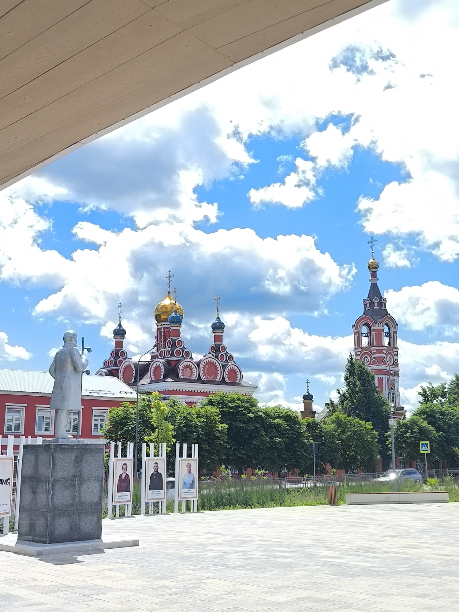 The image captures a view of the Church of the Nativity of the Blessed Virgin Mary in Taldom, Russia, under a bright blue sky with scattered white clouds. In the foreground, a statue of a man stands on a grey pedestal. To the right of the statue, a series of framed portraits are displayed. The church itself is a prominent feature, characterized by its red and white facade, numerous domes, and a tall, slender bell tower with a red and white striped pattern. Lush green trees surround the church. A paved walkway extends across the foreground, with a modern bench and a trash receptacle visible on the right. A dark-colored car is parked on a road in the background, adjacent to a yellow traffic sign with a pedestrian symbol. The overall scene suggests a public square or park area in front of a historical religious site.