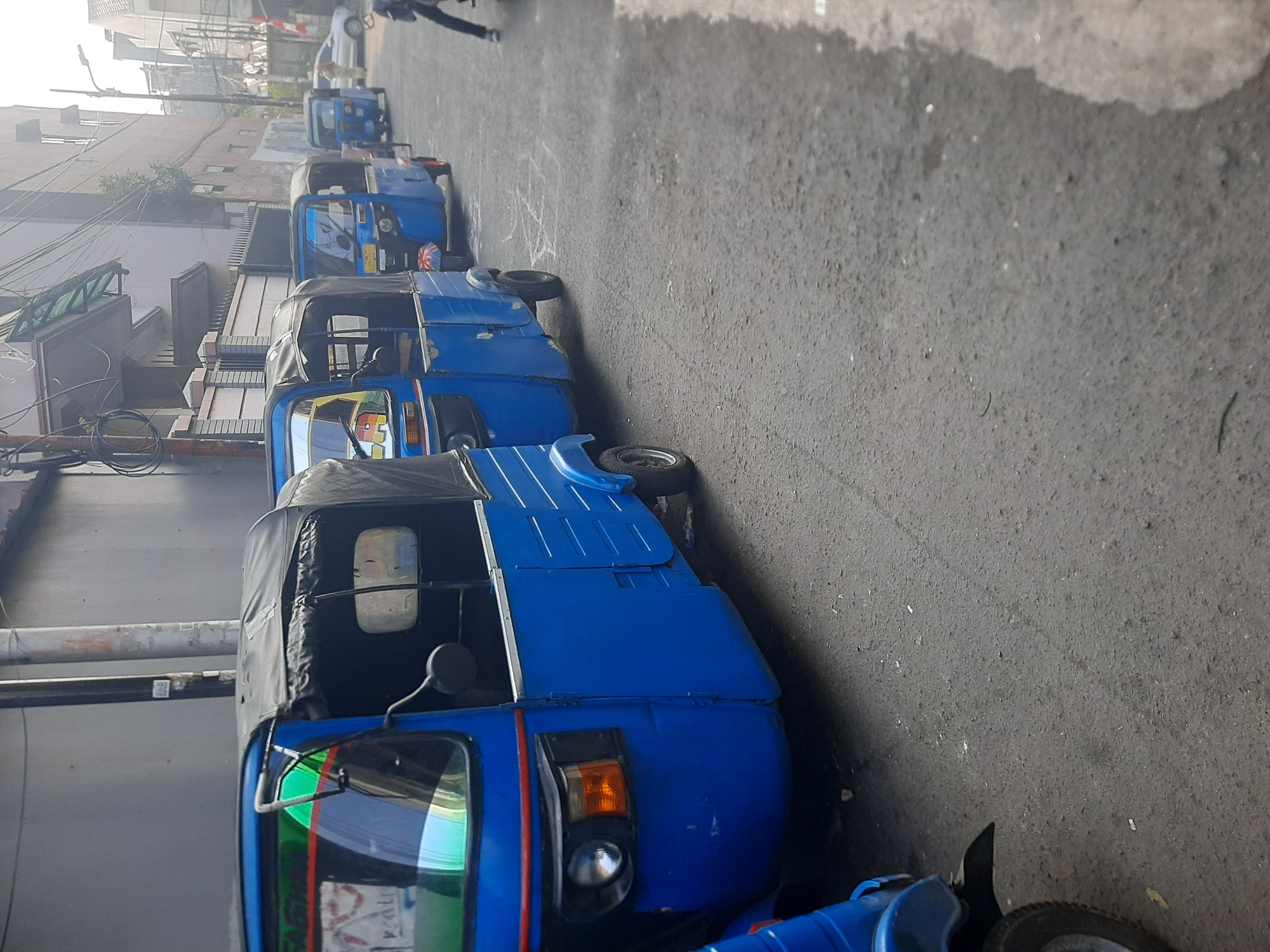 In Central Jakarta, Indonesia, a line of blue auto-rickshaws, commonly known as bajaj, is parked along a paved street. The bajaj are the primary subjects, with their distinct three-wheeled design and canvas canopies. The foreground bajaj is the most prominent, showing its blue body, a black canopy, and a headlight assembly. Behind it, several other bajaj are visible in a receding line, their blue bodies and black canopies creating a repetitive pattern. The street is asphalt and appears somewhat worn. To the left of the bajaj, a building wall with some visible piping and wiring runs along the street. In the background, more buildings and the sky are visible. The lighting suggests it is daytime, possibly morning or late afternoon, given the angle of the shadows. The scene is one of a parked fleet of vehicles in an urban setting.