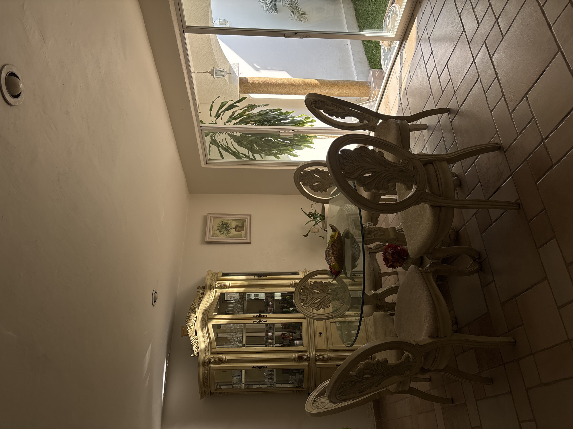 The image captures a dining room interior in an unknown location, possibly Chilpancingo, Mexico. The focal point is a round glass dining table surrounded by ornate cream-colored chairs. A large, decorative china cabinet with glass doors stands against the wall, displaying glassware and bottles. A framed picture hangs above the cabinet. Daylight streams in through a large sliding glass door, revealing a patio with plants and a column outside. The floor is tiled in a terracotta color. No people are visible.