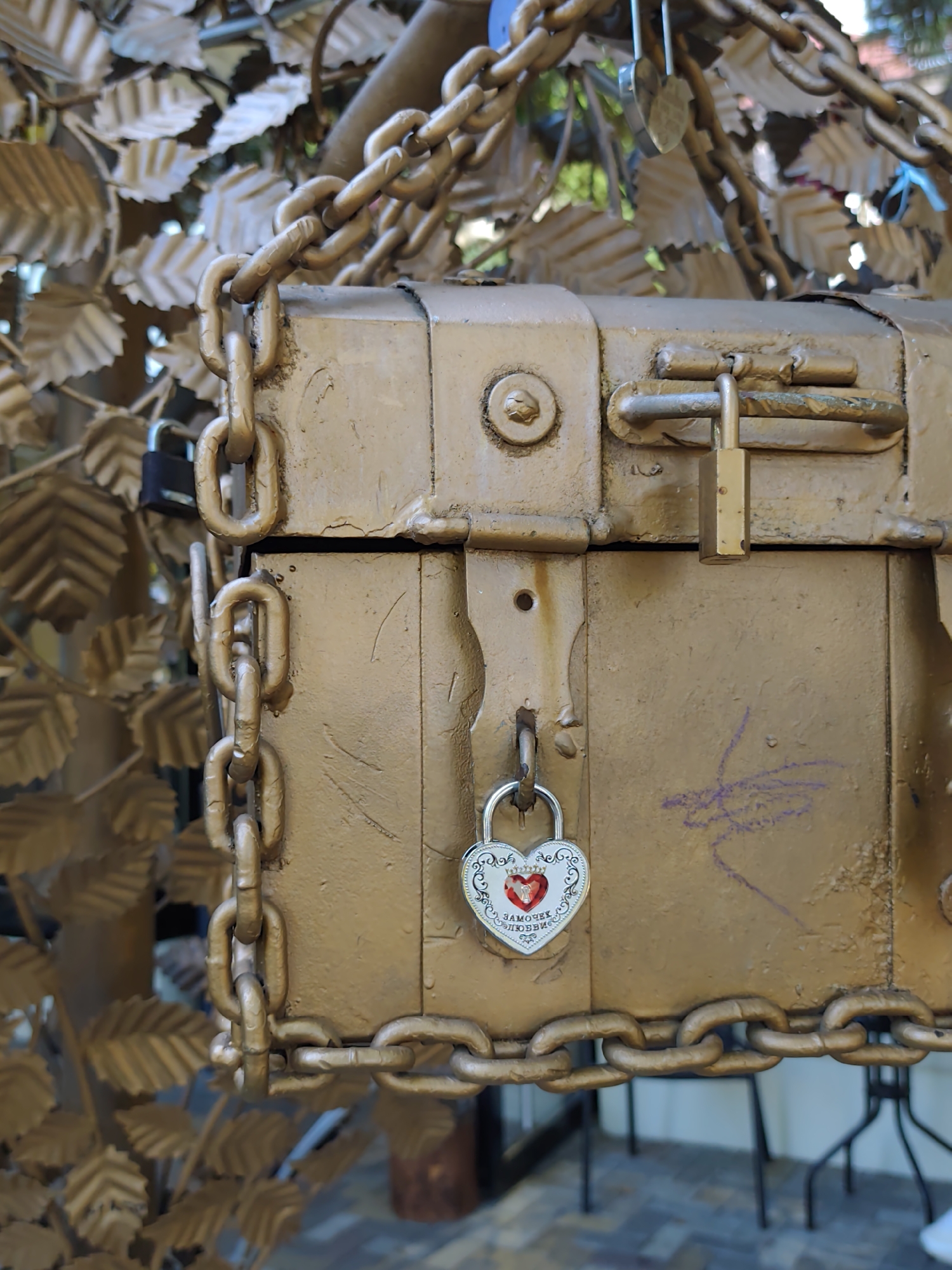 The image shows a close-up of a golden, weathered chest or box secured with chains and locks. The chest is part of a decorative structure that appears to be a gate or fence, adorned with golden leaf-like ornaments. A large chain wraps around the chest and the structure. Two padlocks are visible: a rectangular brass padlock on the main clasp and a heart-shaped silver padlock with red detailing and an inscription hanging from a lower clasp. The inscription on the heart-shaped padlock appears to be in Cyrillic. The background consists of more of the golden leaf decorations and some out-of-focus greenery and structural elements. The scene is likely an outdoor setting, possibly a public space in Sochi, Russia, where love locks are a common tradition.