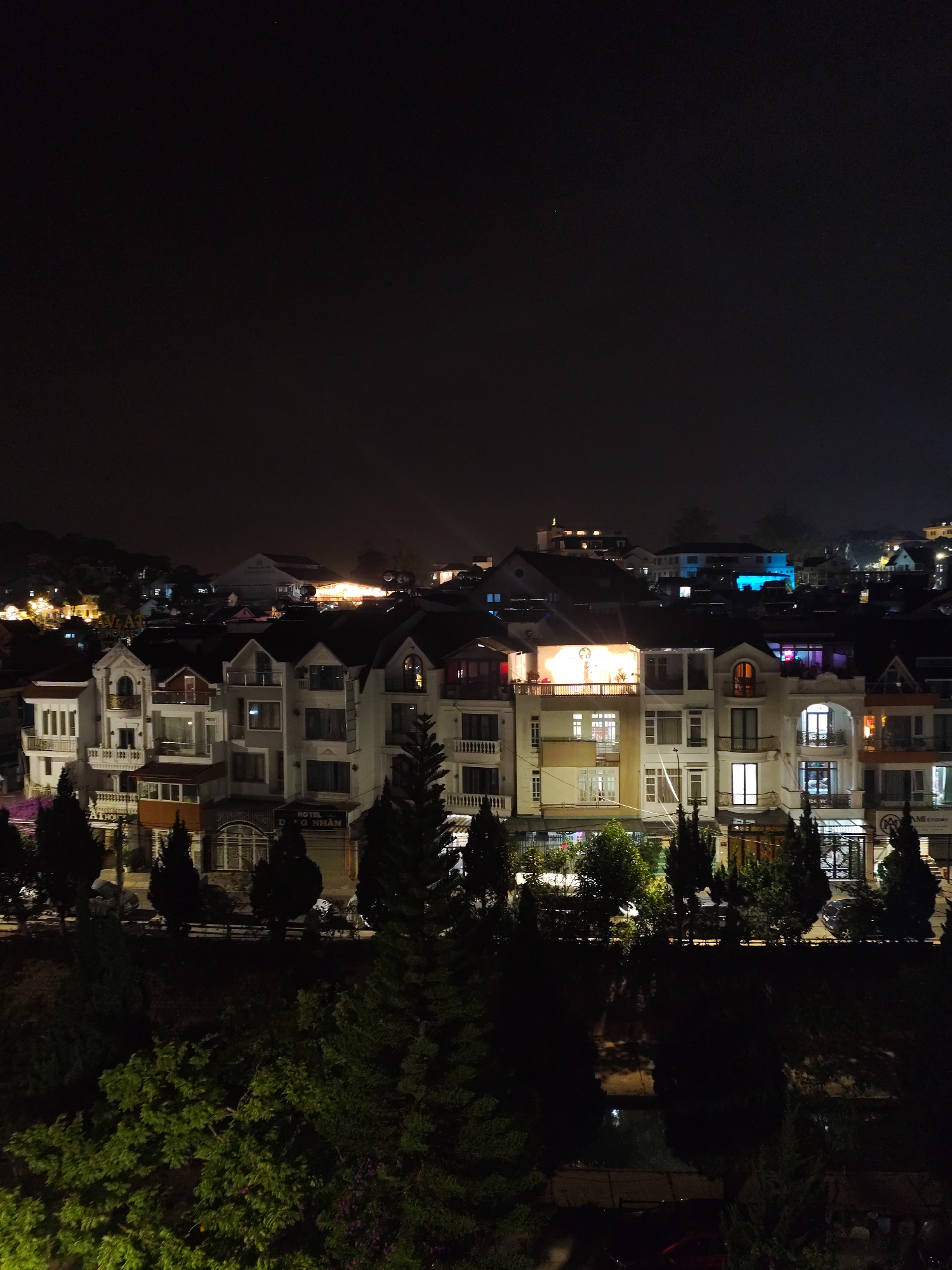 The image depicts a nighttime scene in Da Lat, Vietnam. A row of buildings with varying architectural styles is centrally located in the composition. The structures appear to be multi-story and predominantly white or light-colored, with many visible windows lit from within. Between buildings, there are dark trees and foliage.
In the upper background, further buildings and lights suggest an urban environment extending into the distance. The foreground consists of denser greenery, obscuring the ground level.
The image is taken at night, with a dark sky above, suggesting little ambient light. The illumination comes mainly from the buildings. No people are visible in the image.