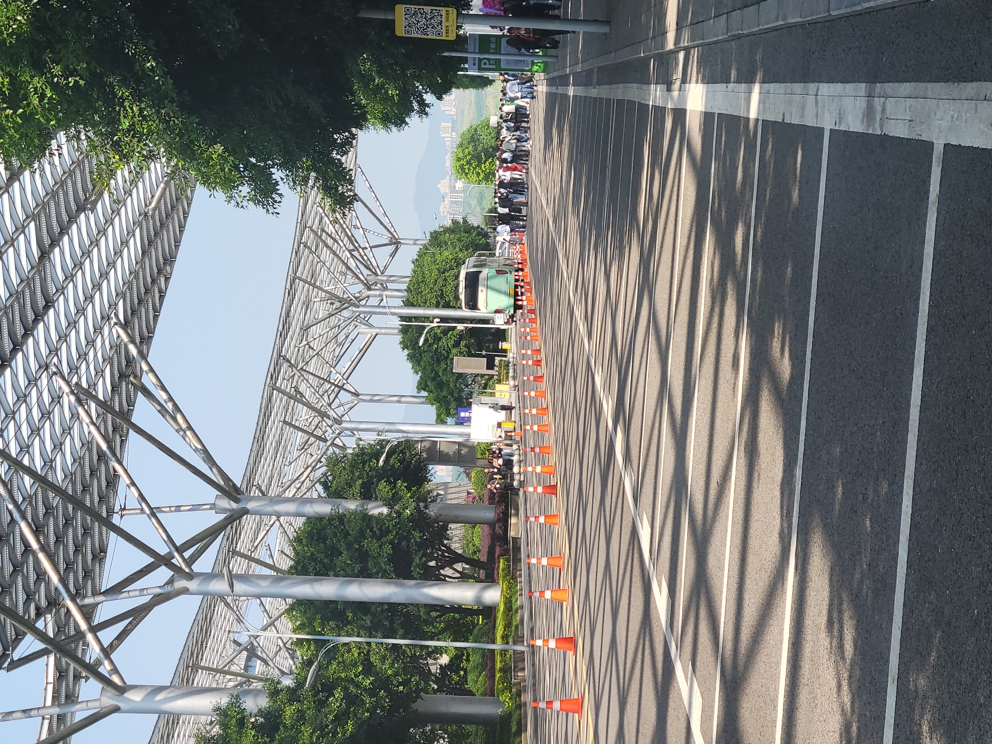 The image depicts a road in the city of Liangjiang, China. The road is marked with white lines and bordered by orange traffic cones. Shadows from an overhead structure cast lines across the road surface. A green bus or tram is visible in the distance. A crowd of people is lined up on the far side of the road. A metal structure runs along the left side of the road, partially covered by trees. A parking sign is visible on the right side of the road.