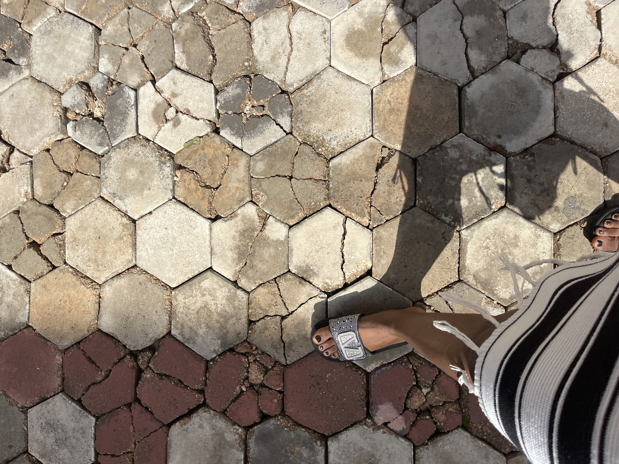 The image shows a person's legs and feet wearing a black sandal with rhinestones and a black and white striped skirt, standing on a pathway made of hexagonal pavers. The pavers are weathered and cracked, with some missing or broken pieces, and they vary in color from reddish-brown to light gray. The setting appears to be outdoors, as indicated by the shadows cast on the ground. The location is specified as Gura Top, Nigeria.
