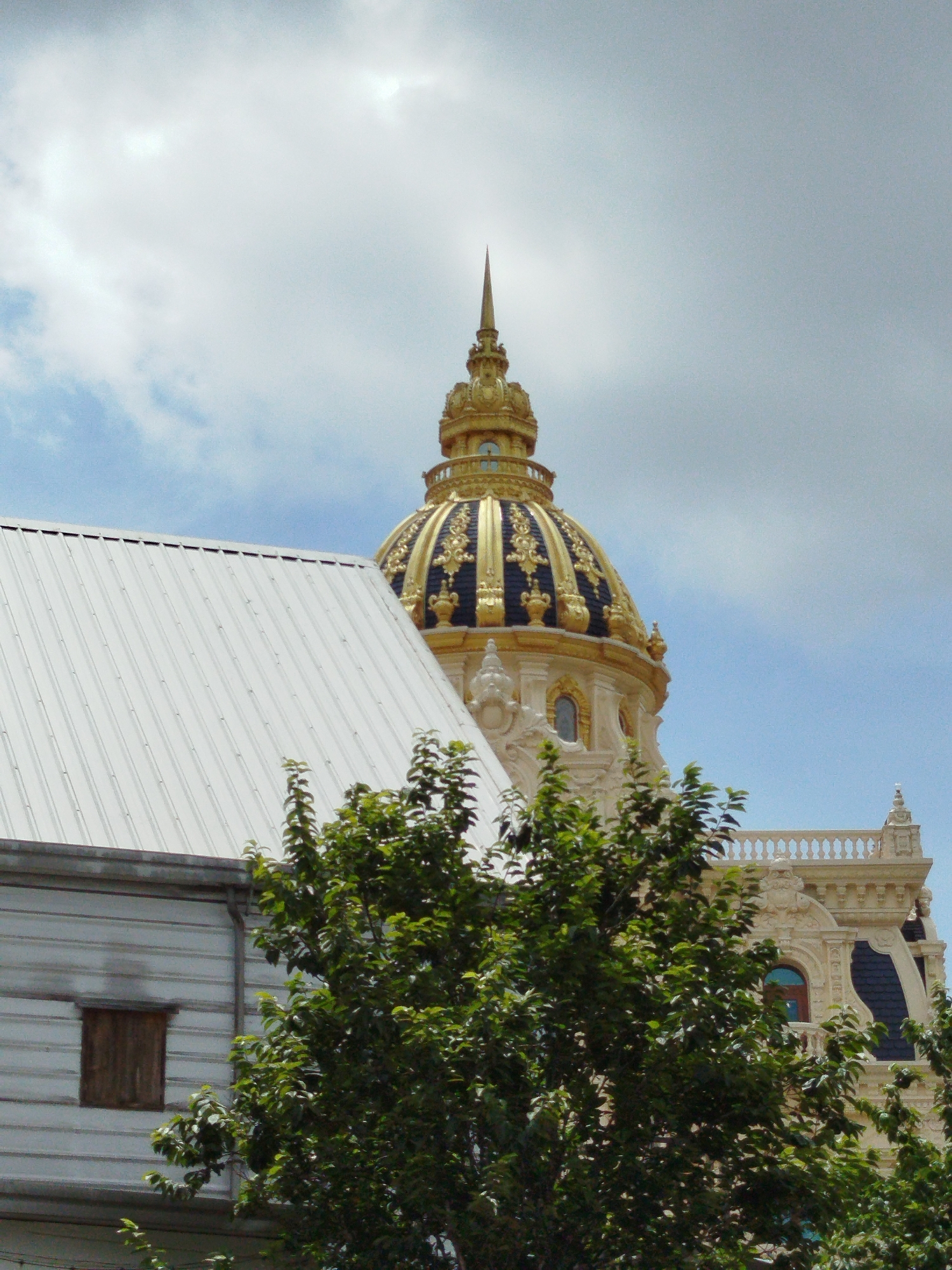 The image captures an exterior view of the Dinh I (also known as the Governor's Palace or Dalat Palace), a historic building located in Da Lat, Vietnam. The primary subject is the distinctive golden dome of the palace, topped with a spire, which is partially obscured by the upper branches of a lush green tree in the foreground. To the left of the dome, a portion of a white, corrugated metal roof of another structure is visible, along with a small, dark wooden window. The Dinh I itself, made of light-colored stone with ornate architectural details, is seen behind the tree, with a balustrade crowning its upper level. The sky above is partly cloudy with patches of blue. The overall scene depicts a sunny day with a mix of modern and historical architecture amidst greenery.