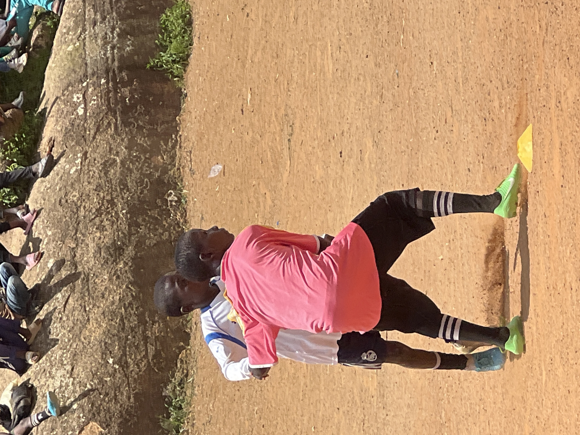 The image shows two young boys playing soccer on a dirt field in Jos, Nigeria. The boy in the foreground is wearing a pink shirt, black shorts, and black and white striped socks, along with bright green soccer cleats. He is facing away from the camera, with his right arm extended. The second boy, partially visible behind him, is wearing a white shirt with blue trim and dark shorts, and light blue soccer cleats. He is looking towards the boy in pink. The background features a large rocky formation, with some sparse green vegetation. The ground is dusty and uneven. A small yellow cone is visible on the ground in the lower right quadrant of the image. The overall scene depicts a casual soccer game or practice in an outdoor environment.