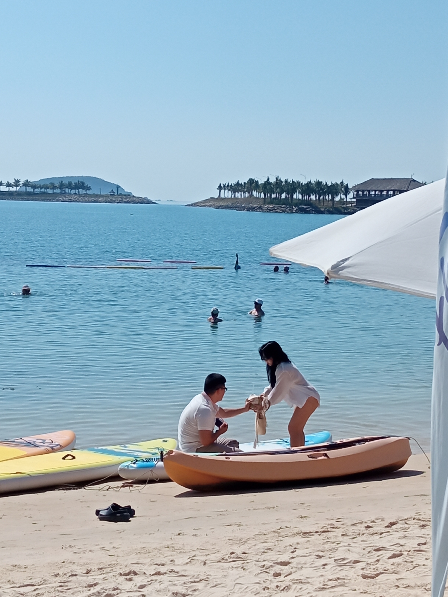 The image depicts a beach scene in Nha Trang, Vietnam, with the Nha Trang Bay in the background. In the foreground, two individuals are interacting with a small boat on the sandy shore. A man, wearing a white polo shirt and khaki pants, is seated in the boat, looking towards a woman. The woman, dressed in a white shirt and pink bottoms, is standing and handing him an object. To the left of the boat, a yellow and a yellow and blue paddleboard are resting on the sand. Further to the left, black sandals are visible on the sand. In the water, several people are swimming, and a line of colorful buoys stretches across the bay. In the distance, islands with palm trees and buildings line the horizon under a clear blue sky. A large white umbrella partially obscures the right side of the frame.