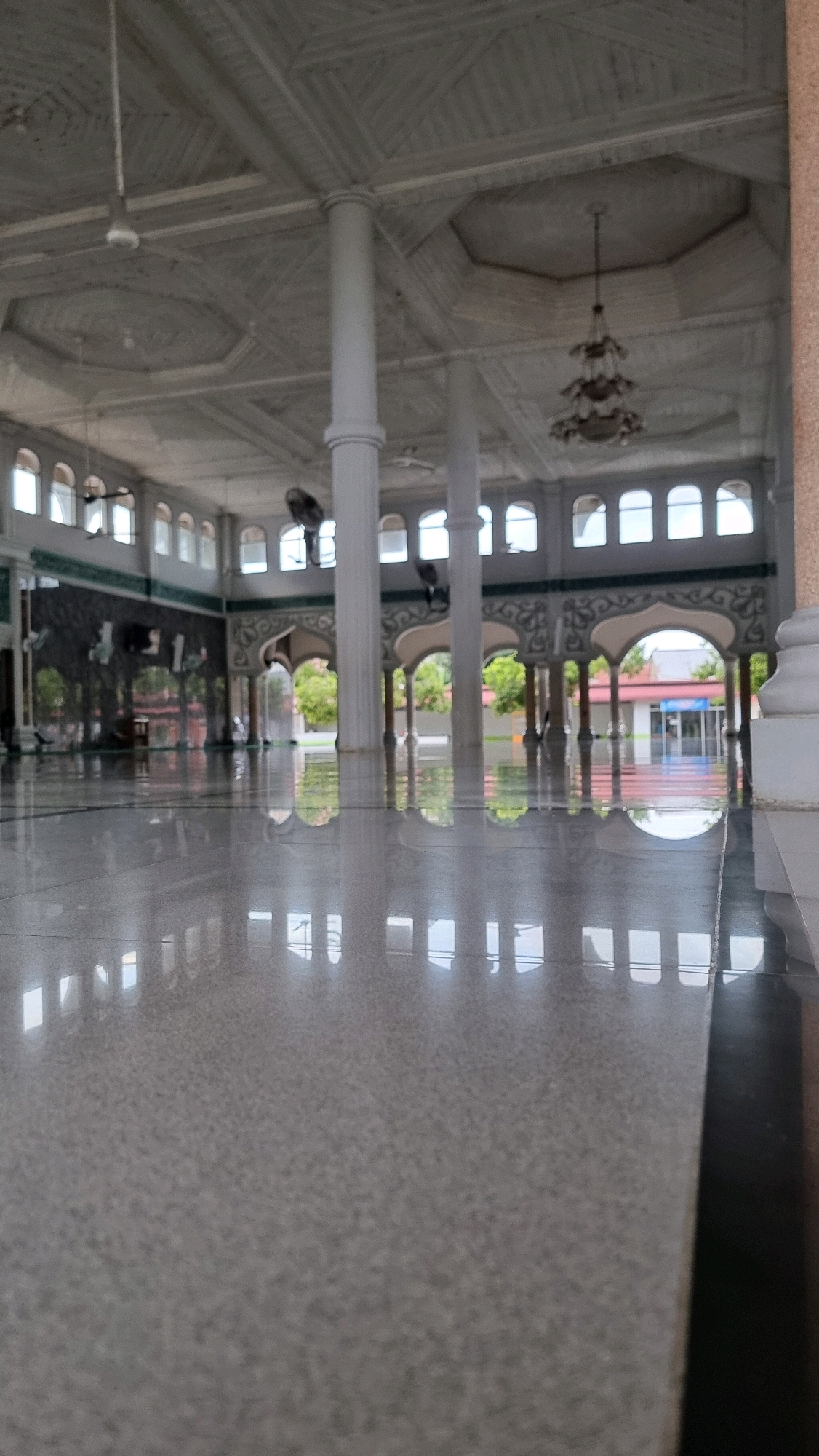 The image depicts the interior of a mosque in Manetunong, Indonesia. The perspective is low, focusing on the highly reflective, speckled granite floor. In the background, a series of arched colonnades support the ceiling. The ceiling itself is intricately detailed with geometric patterns. Light streams in through numerous arched windows, creating reflections on the floor. A chandelier hangs from the ceiling in the mid-ground. The scene appears empty of people. The overall impression is one of architectural grandeur and tranquility.