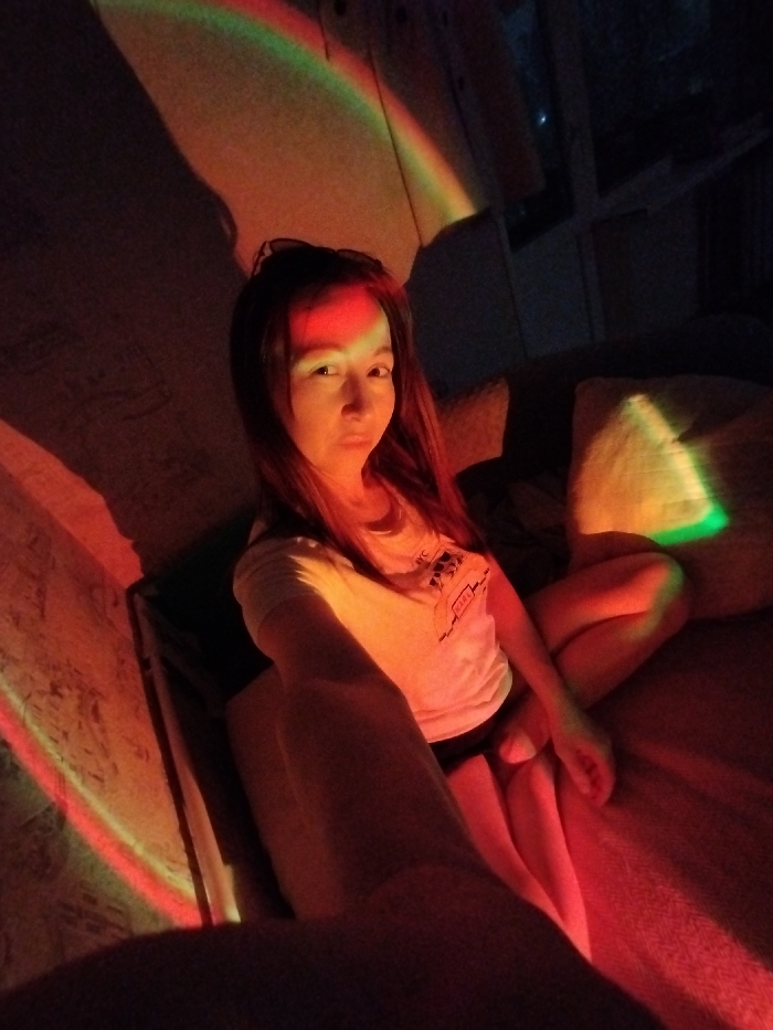A woman sits cross-legged on a red-lit surface, possibly a bed. She wears a white t-shirt and dark shorts. A rainbow-colored light source projects onto her face, the wall behind her, and a pillow to her right. The lighting is dramatic, casting strong shadows. She looks directly at the camera with a slight pout. Sunglasses are perched atop her head. The overall setting appears to be indoors.