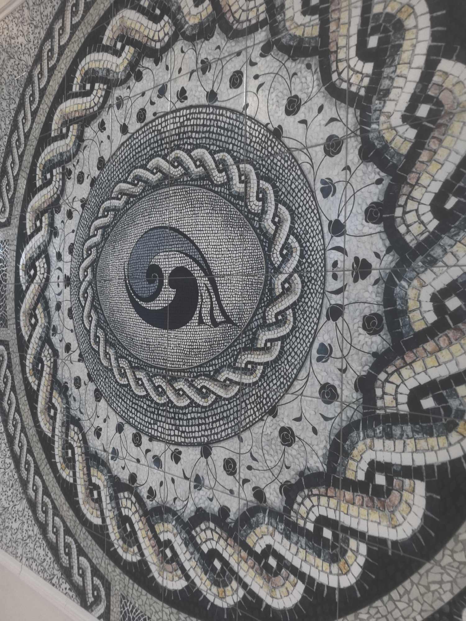 The image shows a mosaic on a wall, featuring an oval design with concentric bands. The central element is a spiral pattern in shades of gray and black. Surrounding this are bands of intricate mosaic work: a densely patterned band, a wavy chain-like design, a background with floral motifs, and an outer band of interconnected shapes in black, white, and beige tiles. The overall impression is one of craftsmanship and artistic detail. This mosaic is located in the city of Nizhny Novgorod, Russia.
