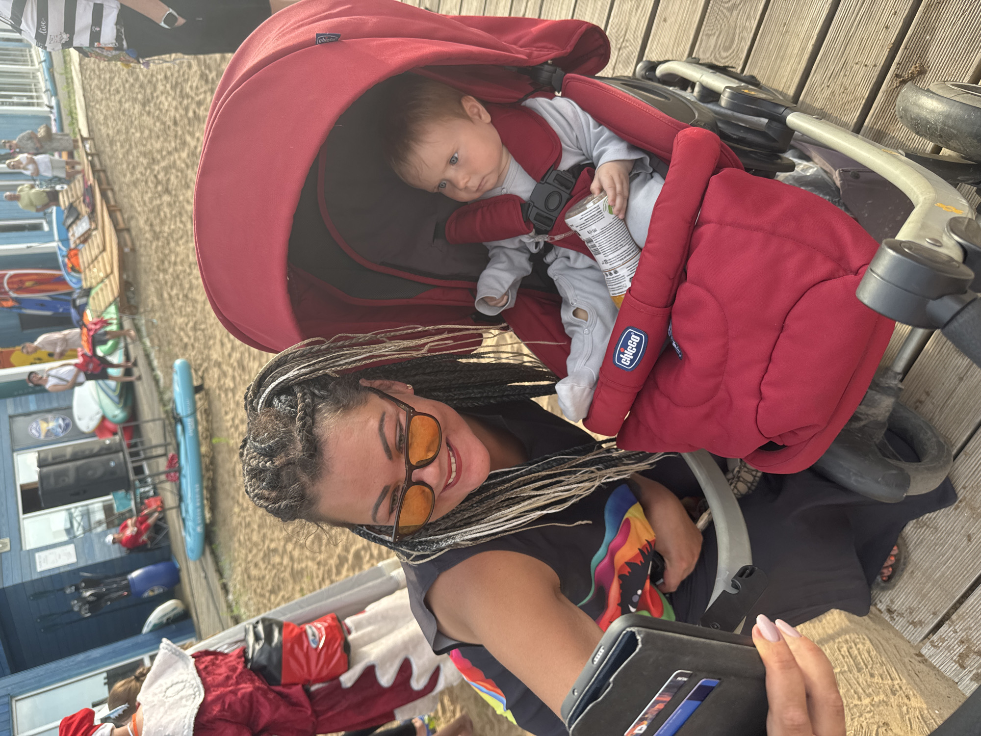 A woman with braided hair and sunglasses is taking a selfie with her baby in a stroller. The baby is wearing a grey outfit and holding a bottle. The stroller is red and has the Chicco logo. The scene is set on a sandy beach with a wooden boardwalk. In the background, there are people, a building with a blue facade, and various water sports equipment. The overall setting appears to be a recreational area in Chaikovsky, Russia, likely a beach resort or park.