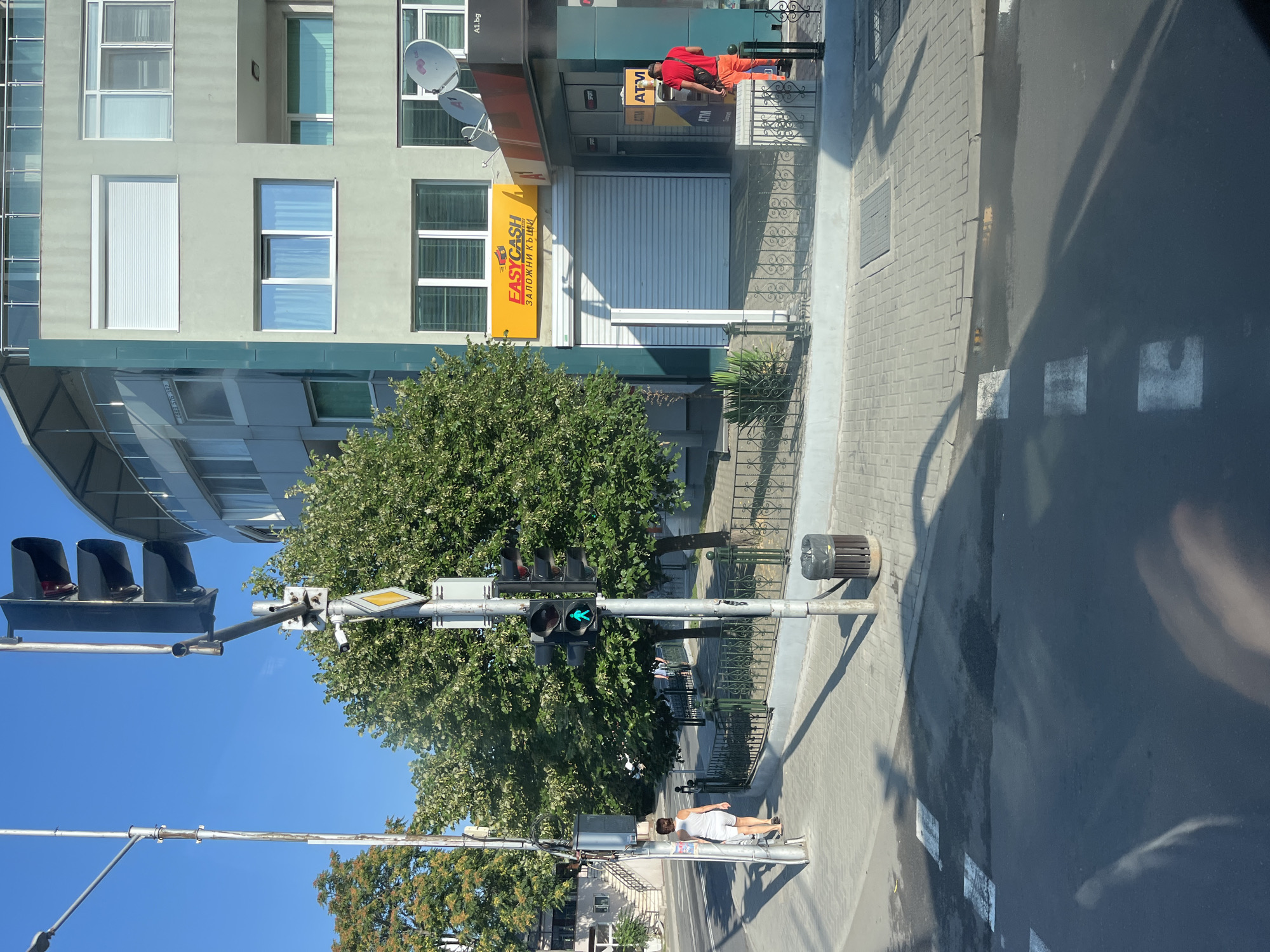 The image captures a street scene in Nesebar, Bulgaria, under a clear blue sky. A traffic light prominently displays a green pedestrian signal, indicating it is safe to cross. To the left, a woman in a white tank top and shorts walks away from the camera on a paved sidewalk. Behind her, a tall lamppost stands.
The background features a modern, multi-story building with a green facade. Several windows are visible, some with closed blinds and others with sheer curtains. A prominent yellow sign with "EASYCASH" and Bulgarian text is mounted on the building. Adjacent to this is an entrance with ATMs, and a man in a red shirt and orange pants is seen standing near one of the ATMs. Trees with lush green foliage border the sidewalk and line the street. A metal fence with decorative elements separates the sidewalk from the building. A trash receptacle is visible near the traffic light pole. The road in the foreground has a wet appearance, possibly due to recent rain, with white pedestrian crossing markings. Shadows from the traffic light and a person are cast across the road and sidewalk.