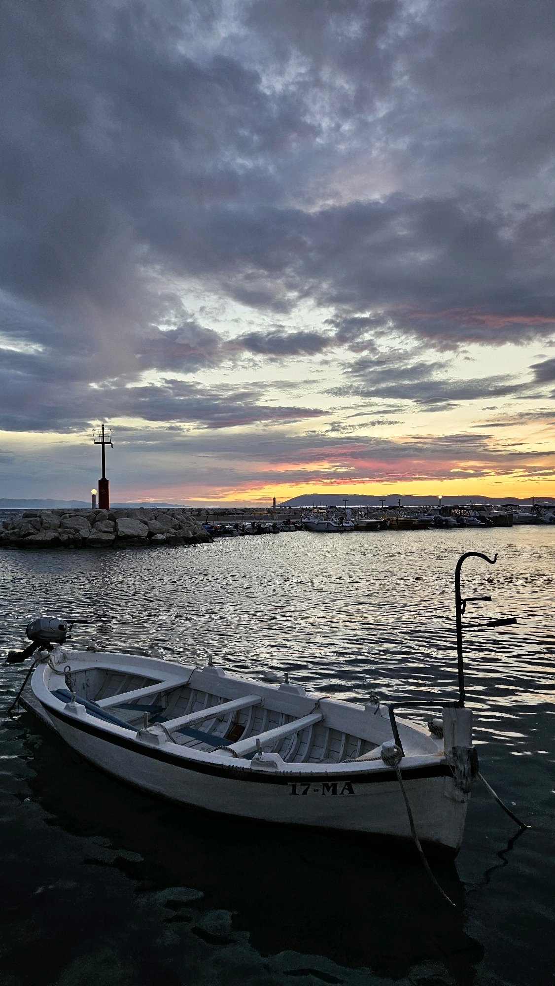 This is a vertical, eye-level, medium shot of a small white boat floating on dark, rippling water at sunset. The boat is positioned in the lower-left quadrant of the frame and is facing towards the right. It is an open vessel with wooden seating and a small outboard motor attached to the stern. The name "17-MA" is visible on the side of the boat.
In the background, a harbor breakwater made of large rocks extends from the left into the water. A red navigation light with a smaller yellow light below it is mounted on a pole at the end of the breakwater. Beyond the breakwater, several other boats are docked, and a faint silhouette of hills or mountains is visible on the horizon.
The sky above is filled with dramatic, cloudy formations, with bands of pink, orange, and yellow hues indicating the setting sun. The clouds are a mixture of dark grays and lighter, wispy whites and purples.
The scene appears to be a harbor in Općina Tučepi, Croatia, based on the visual cues of a coastal town with a marina and Mediterranean-style hills in the distance. The overall atmosphere is calm and serene, characteristic of a peaceful evening by the sea. There are no people visible in the image.