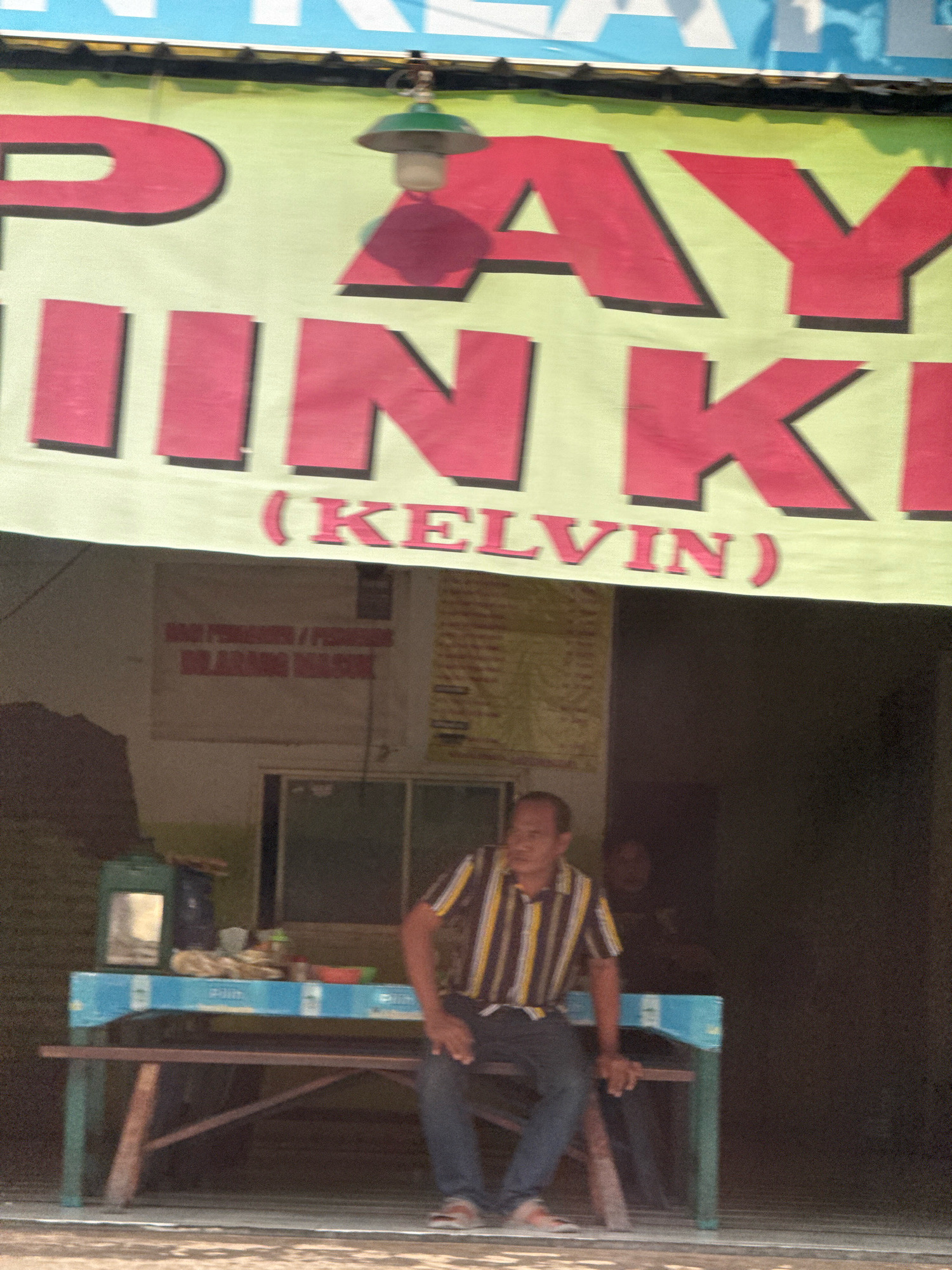 The image depicts a street scene in Surabaya, Indonesia. In the foreground, a man wearing a striped shirt and jeans is seated on a bench in front of a stall. The stall has a blue tabletop and is laden with various items, including containers and possibly food. Behind the man and the stall, a banner with large, red lettering is displayed. The banner appears to say "PAY JIN KI (KELVIN)". The lettering is in a bold, sans-serif font with a dark shadow. A green lamp with a bare bulb hangs from the banner. In the background, a building with some signs is visible, though the text on these signs is mostly illegible. The overall impression is that of a casual, everyday scene in a local commercial area.