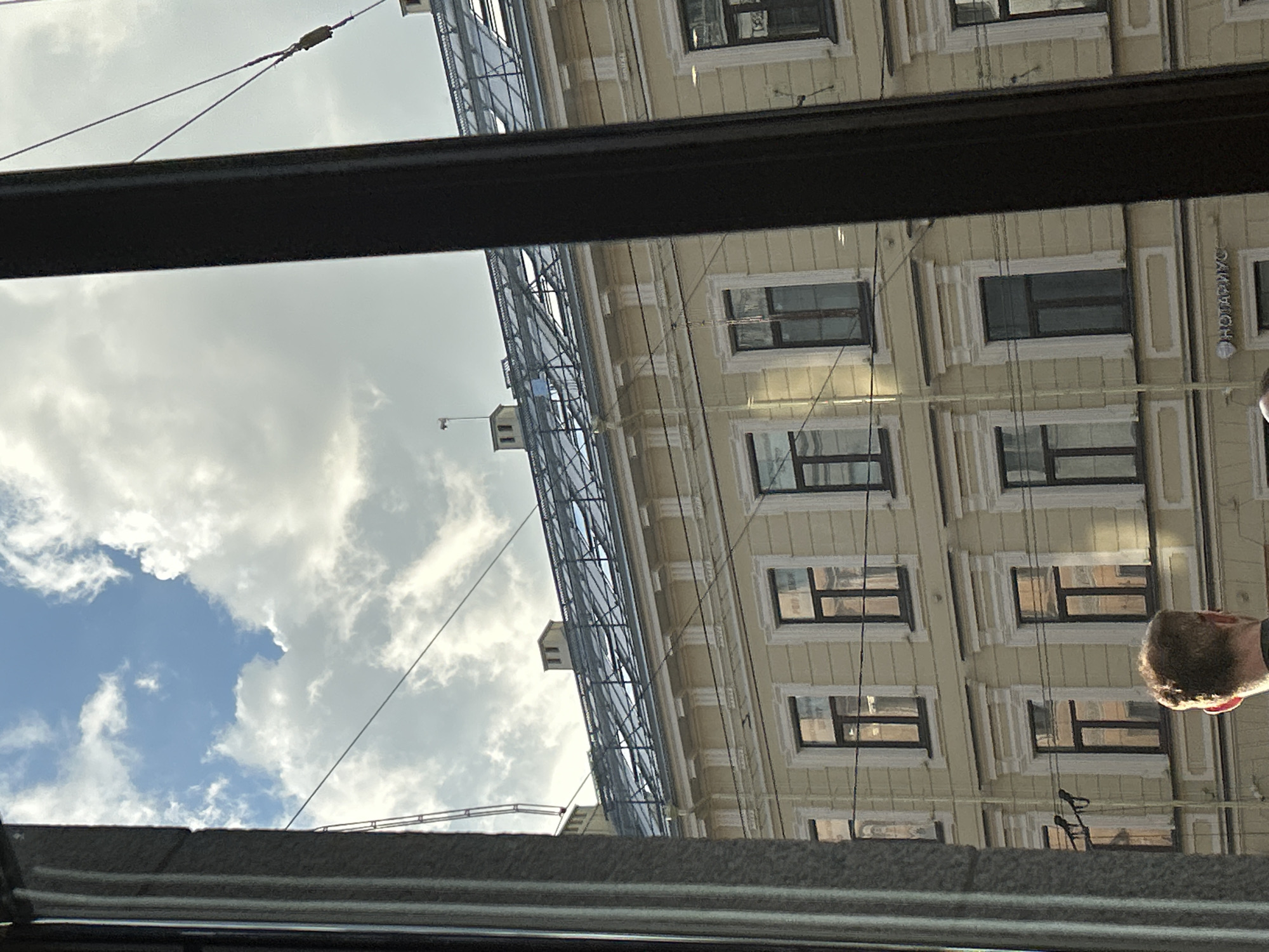 The image, taken from inside a building and looking out a window, captures a section of a pale yellow, multi-story building's facade. The building features regularly spaced dark-framed windows, some with ornate white trim. Above the windows, a decorative frieze runs horizontally. The roofline is visible, with what appears to be a glass-covered attic or conservatory structure. The sky above is partly cloudy, with a mix of blue sky and white clouds illuminated by sunlight. Several electrical wires crisscross the scene, casting shadows. In the lower left portion of the image, a person's head and shoulders are visible from behind, with light brown hair. In the lower right, a dark vertical structural element, likely a window frame or pillar, partially obscures the view.