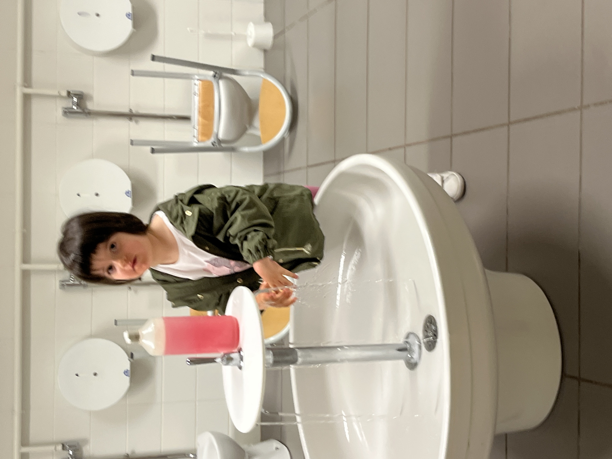 The image captures a young child, likely a girl with dark, bob-cut hair, washing her hands at a sink. She is dressed in a dark green jacket over a white t-shirt and looks directly at the camera with a neutral expression. Water is actively running from a tall chrome faucet into the white, wall-mounted sink, splashing onto her hands. A large bottle of pink liquid soap with a pump dispenser rests on a small, round white shelf attached to the faucet's base.
The setting appears to be an indoor public restroom, possibly in a facility designed for children, such as a school or daycare, indicated by the low height of the sink. In the background, an inverted metal and light wood stepping stool leans against the wall, likely used for children to reach the fixtures. The walls are covered in white rectangular tiles, and the floor is composed of light gray tiles. Several white, circular wall-mounted dispensers, which could be for paper towels or toilet paper, are visible on the tiled wall.
The activity taking place is the child hand-washing. There are no specific time-of-day or weather cues, as the scene is indoors. No visible text is present in the image. The location context is La Fouillouse, France.