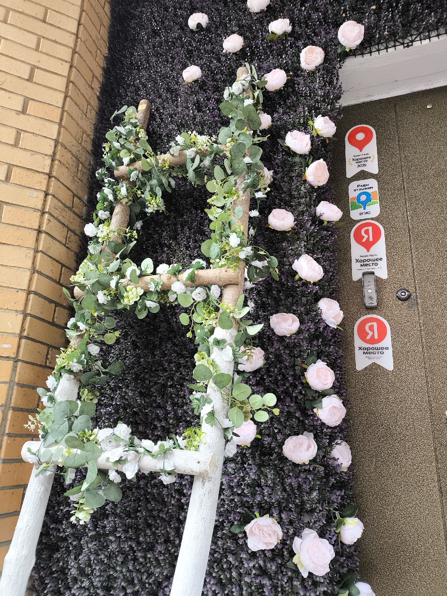 Here's a detailed description of the image:
**Scene:** The image shows an exterior decoration next to a brick building. The decoration consists of a wooden ladder adorned with white and green foliage, positioned against a wall covered in a dark green/purple artificial plant texture. Scattered across this textured wall are light pink peony-like flowers.
**Objects:**
* **Ladder:** A wooden ladder painted white with natural wood showing through, wrapped with green and white artificial foliage.
* **Wall covering:** A dense artificial plant wall covering of dark green/purple color.
* **Flowers:** Artificial light pink flowers, likely peonies, are affixed to the wall covering.
* **Signs:** Several signs are visible to the right of the floral decoration, affixed to what seems to be a door. These signs are in Russian and appear to be related to location services/recommendations. The Yandex logo is visible.
**Setting:** The scene appears to be an entrance to a business or establishment.
**Environment:** The scene is outdoors, next to a brick building with tan bricks.
**Moscow, Russia:** Since the signs prominently feature the Yandex logo (a major Russian tech company) and are written in Russian, it's reasonable to assume the location is likely in Russia. Specifically, Moscow is a plausible location, as Yandex has a strong presence there.