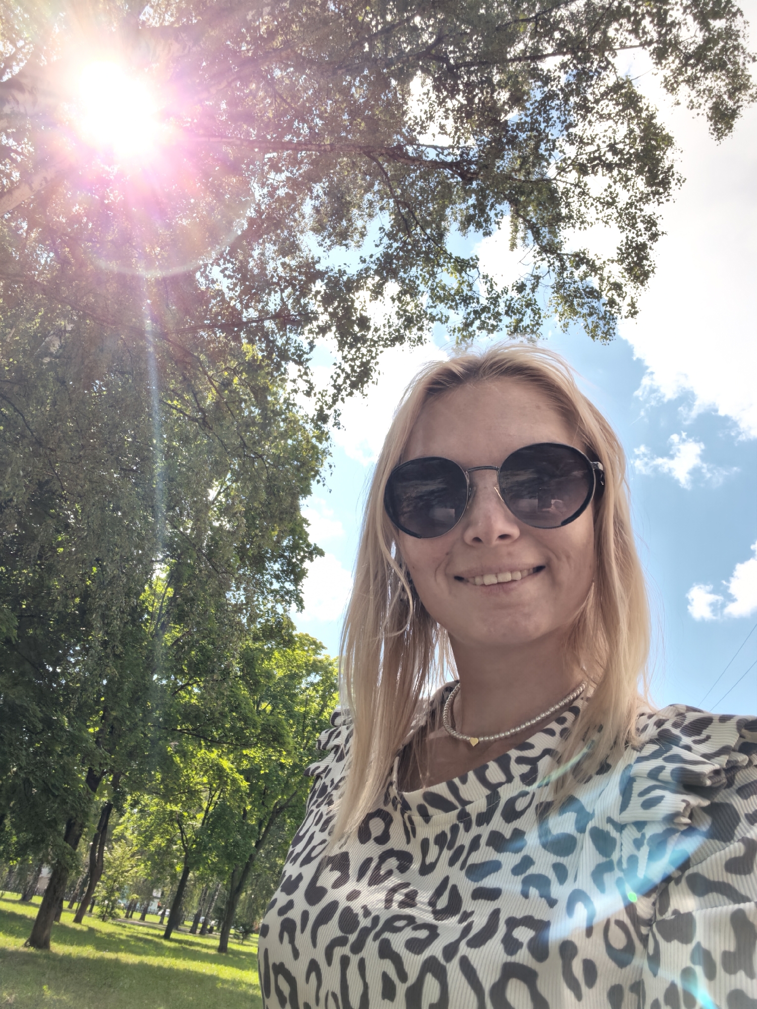 A young blonde woman wearing round sunglasses and a leopard-print shirt is taking a selfie outdoors. She is smiling and her head is tilted slightly. She is wearing a pearl necklace with a small gold heart charm. The background shows a park with green grass, trees, and a blue sky with white clouds. The sun is shining brightly, causing lens flare. The scene appears to be in a park in the city of Sterlitamak, Russia, on a sunny day.
