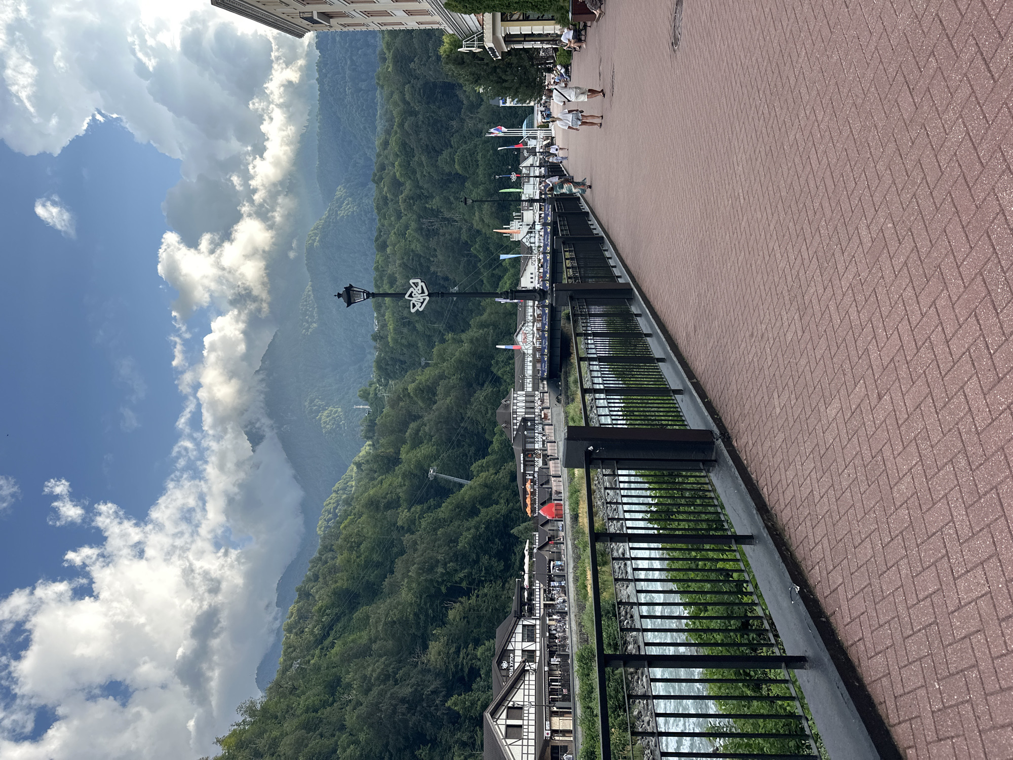 The image shows a picturesque scene in the city of Estosadok, Russia, featuring a cobblestone pathway lined with a black metal fence. A river flows alongside the pathway, its water appearing rough and rapid. Beyond the river, several buildings are visible, some with traditional German-style architecture, and beyond that, lush green mountains rise majestically into a partly cloudy sky. A lamppost stands tall on the pathway, and a few people are strolling along, enjoying the scenic view. The overall atmosphere is tranquil and inviting.