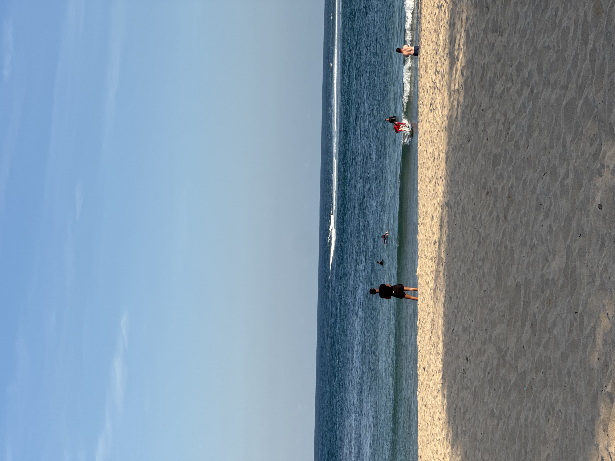 A wide shot of Kuta Selatan, Indonesia, shows a sandy beach leading to the ocean under a clear blue sky. The foreground is dominated by the textured sand of the beach, with gentle waves lapping at the shore. Several people are visible in the water. In the middle ground, a lone figure stands facing the ocean, dressed in black shorts and a black t-shirt. Further out in the water, other individuals are engaged in activities, including what appears to be surfing. A jet ski leaves a white wake in the distance. The ocean is a deep blue, with white crests on the waves. The sky is a gradient of light blue, with subtle wisps of clouds near the horizon.