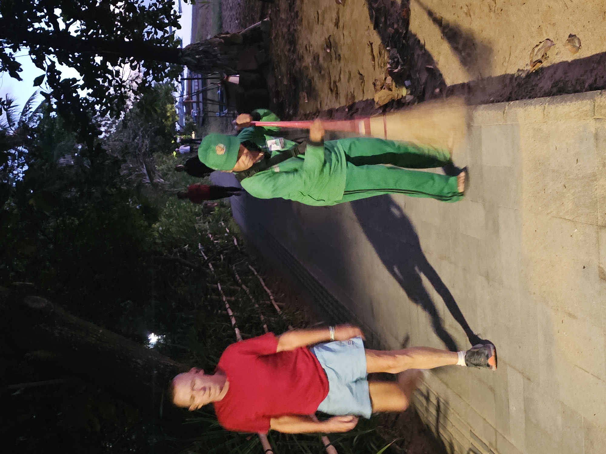 The image shows two people in an outdoor setting. One person, wearing a red shirt and light blue shorts, stands on a paved walkway near a grassy area and trees. Another person, dressed in a green tracksuit and cap, sweeps the walkway. The setting appears to be a park or public space in Denpasar, Indonesia, suggested by the trees and vegetation typical of a tropical climate. The image seems to be taken at dusk or dawn, based on the ambient light.