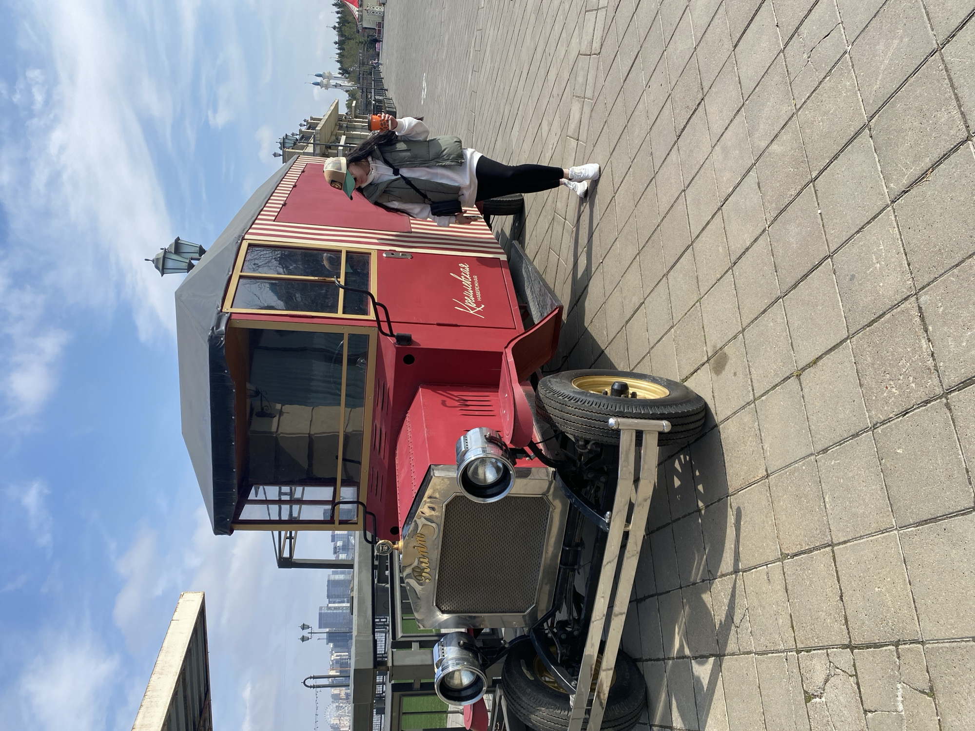 A woman stands next to a red and white striped vehicle, possibly a replica vintage bus or car. The woman is wearing a white sweatshirt, a dark green vest, black leggings, and white sneakers. She also has a baseball cap and is holding a cup. The vehicle has a brass radiator with the name "Кремлевская" written in Cyrillic, which translates to "Kremlin". The location appears to be in Kazan, Russia, based on the architecture and Cyrillic text. The background shows a paved area with some greenery and buildings in the distance. The sky is blue with scattered clouds.