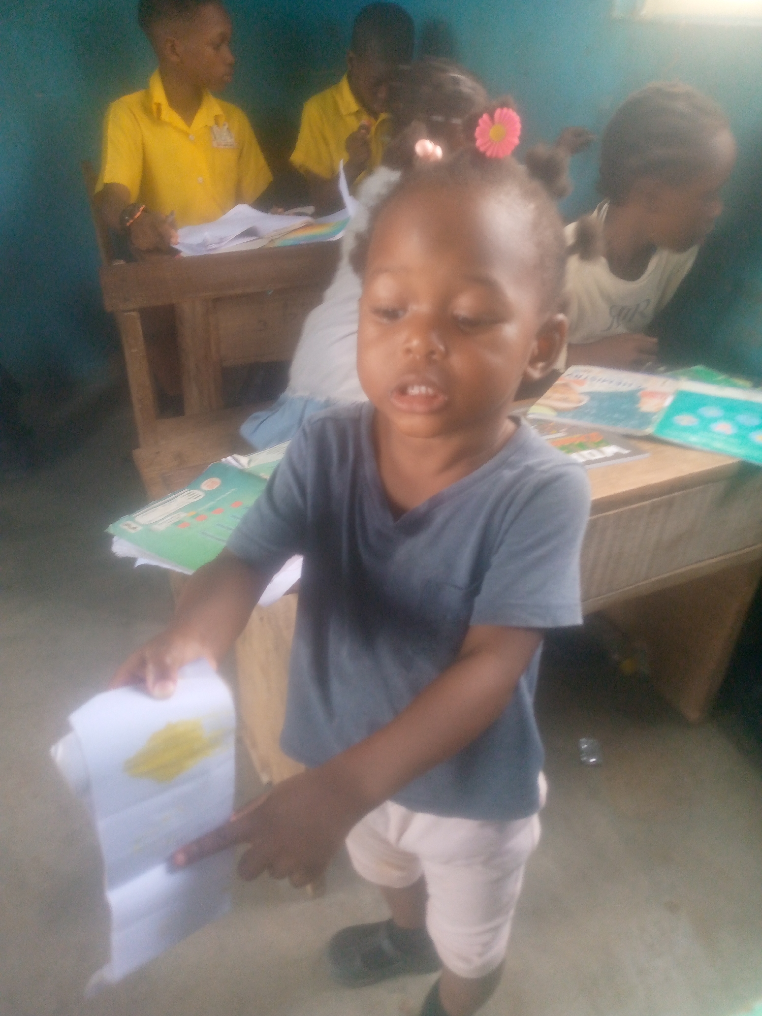 The image shows a young Black child standing in what appears to be a classroom or educational setting. The child is wearing a gray t-shirt, light pink shorts, and black shoes. They are holding a piece of paper with a yellow stain on it and pointing to the paper with their right index finger. Other children are seated at desks in the background. Two are wearing yellow shirts and are working on papers at wooden desks. Another child is visible behind a wooden desk, near a textbook. The location is possibly a school or learning center in Ikorodu, Nigeria, based on the subjects and environment.