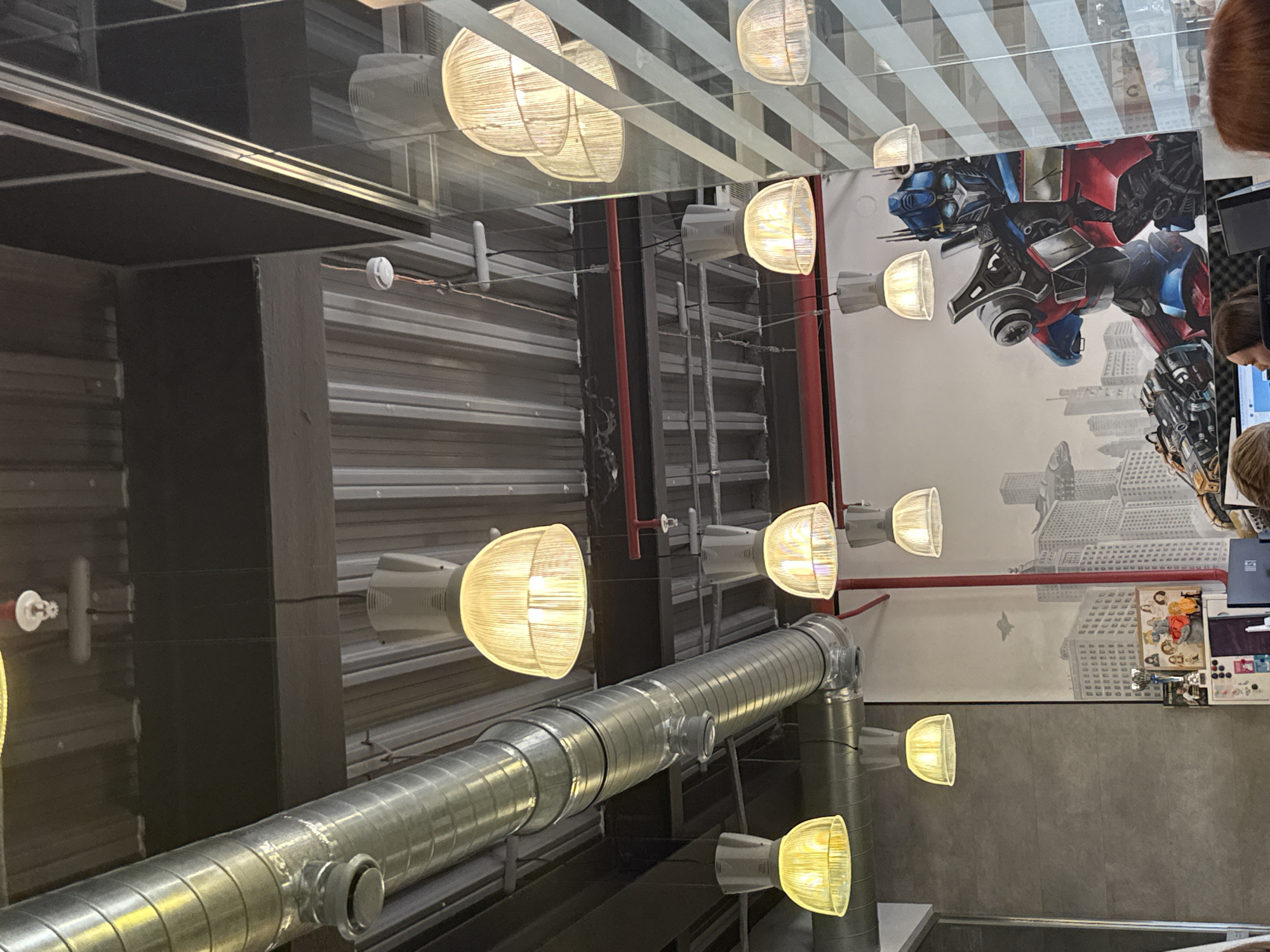 The image depicts an indoor scene featuring an office or commercial space with a modern industrial design. The ceiling is a corrugated metal structure, with exposed industrial ductwork and a prominent metal ventilation pipe running horizontally across the frame. Numerous pendant light fixtures with translucent ribbed shades hang from the ceiling, emitting a warm yellow light.
On the wall, there is a large mural of Optimus Prime, a character from the Transformers franchise, in a dynamic pose. The mural is set against a backdrop of a cityscape that appears to be inspired by New York City, with tall buildings and an aircraft in the sky. Below the mural, a counter or desk area is visible, displaying various items including a trophy, framed pictures, and what appears to be a computer monitor.
Several individuals are partially visible in the foreground, suggesting an active workspace. Their faces are not clearly discernible. The overall environment appears to be well-lit due to the multiple light fixtures. There is a large pane of glass on the right side of the frame, reflecting the interior and the lights. No specific landmarks or features definitively place this location in the city of Perm, Russia.