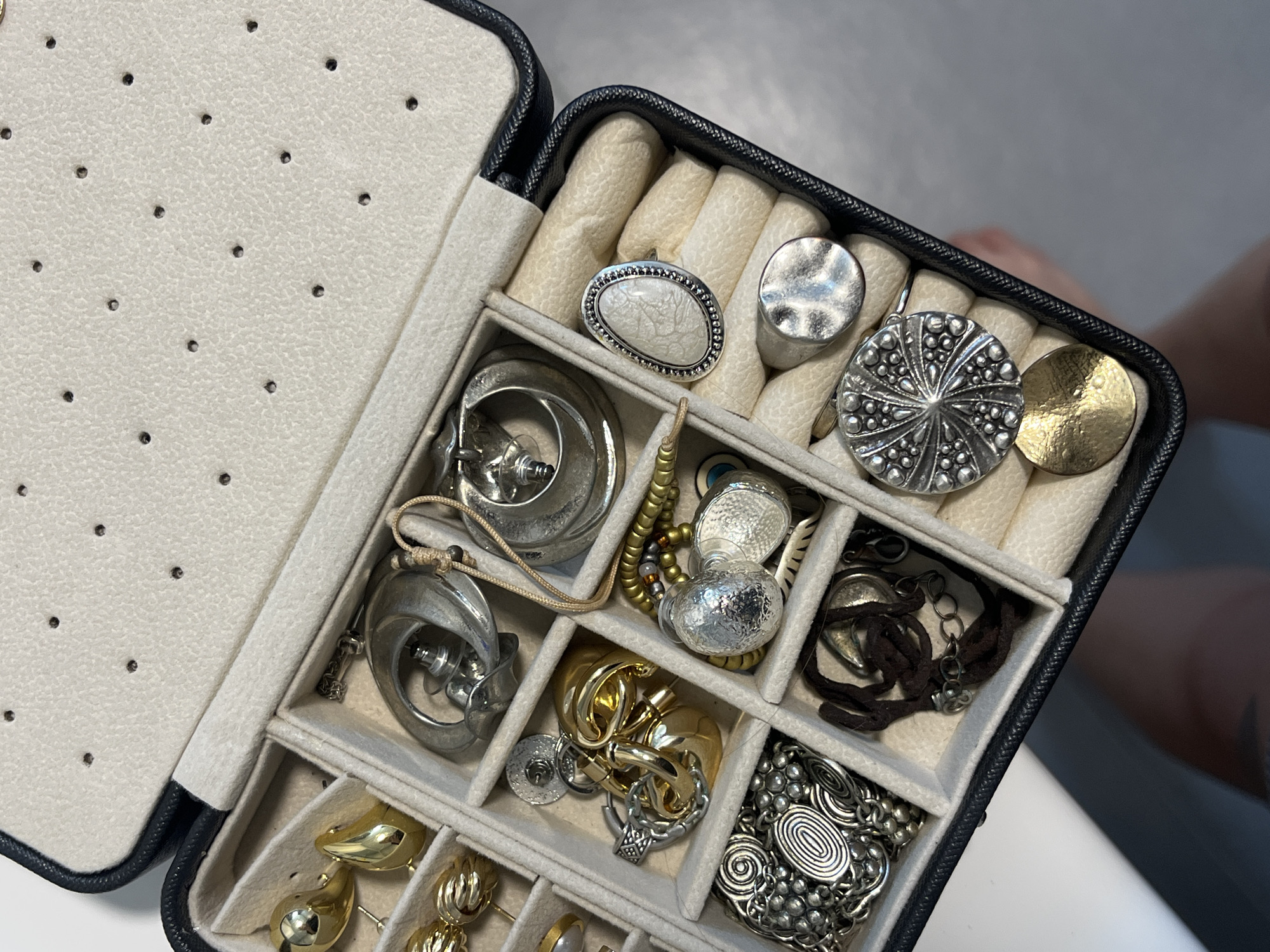 A partially open black jewelry box, lined with off-white fabric, rests on a white surface. The box's interior is divided into compartments, holding various pieces of jewelry, including earrings, rings, and necklaces. The jewelry is predominantly silver and gold in color, with diverse designs and textures. The inside lid of the box has a series of small, evenly spaced holes. The scene is well-lit. No specific location in Limassol, Cyprus, is discernable from the image. No people are fully visible.