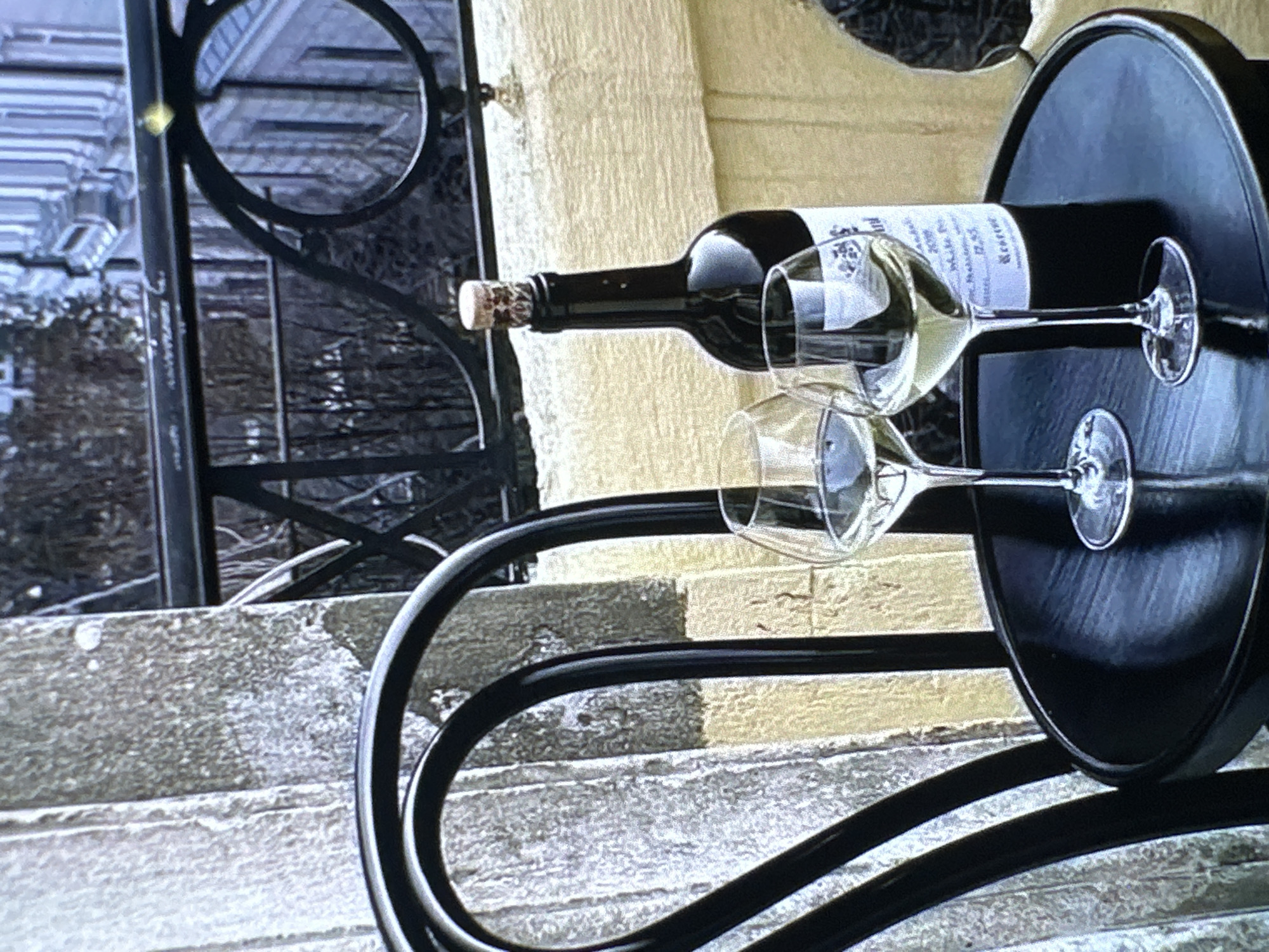 A still life shows a horizontal black wine bottle with a cork in place, resting on a black round metal side table, accompanied by two empty wine glasses of similar shape. The wine bottle has a white label that is only partially legible. The table has decorative black metal legs. The table rests on concrete steps with a weathered appearance. In the background, a railing and a building façade are visible, hinting at an outdoor setting. Without additional information, it's not possible to confirm this is in Yerevan, Armenia.