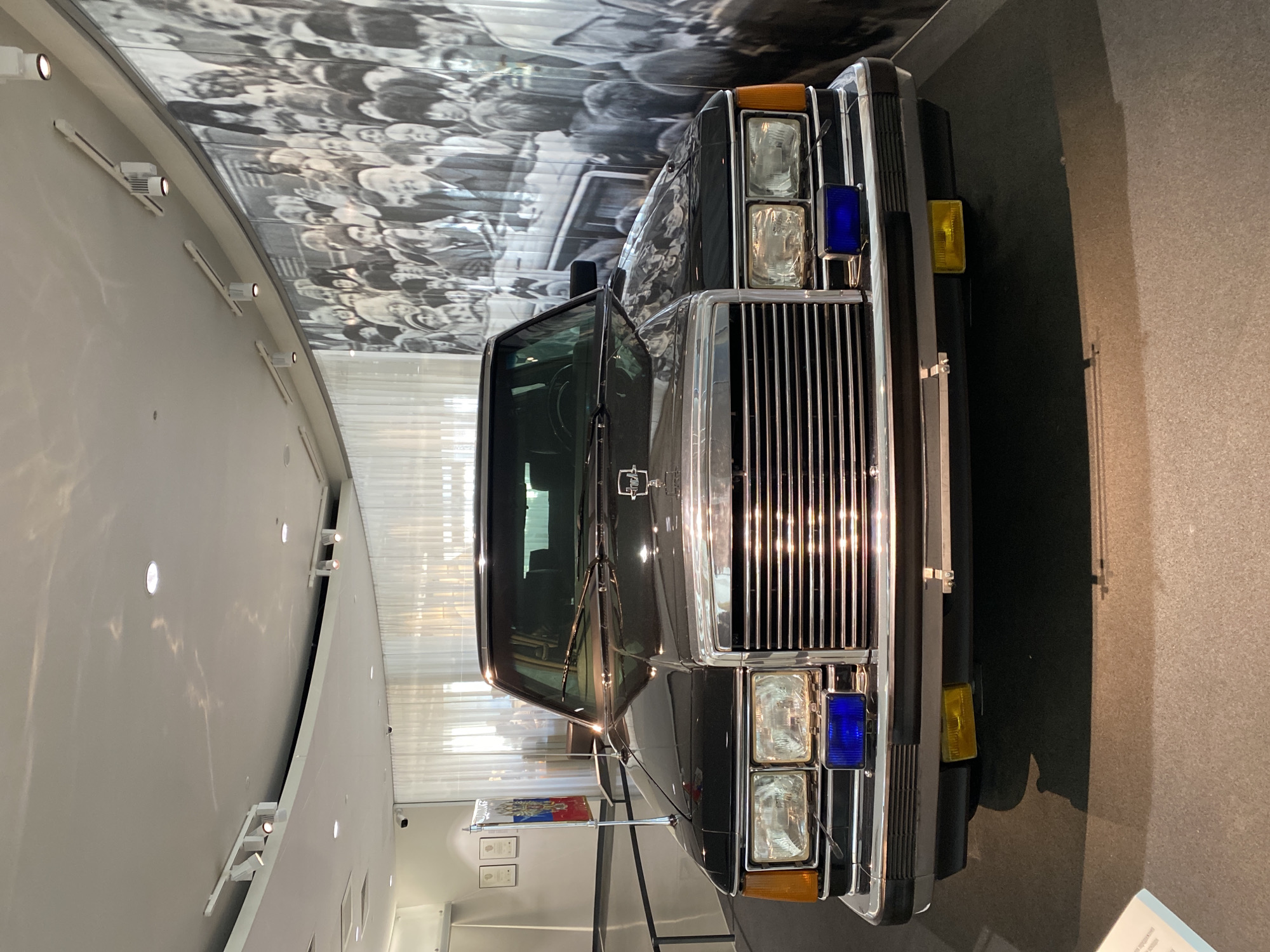 The image shows the front of a black ZIL-41047 Soviet limousine displayed in a museum. The car has a large chrome grille, square headlights with blue and amber lights, and is positioned against a backdrop of a grayscale mural depicting a crowd of people. The car is located in the Yeltsin Center museum in Yekaterinburg, Russia. The floor is grey, and a wall with track lighting is visible in the background. A small display with the Russian flag is next to the car.