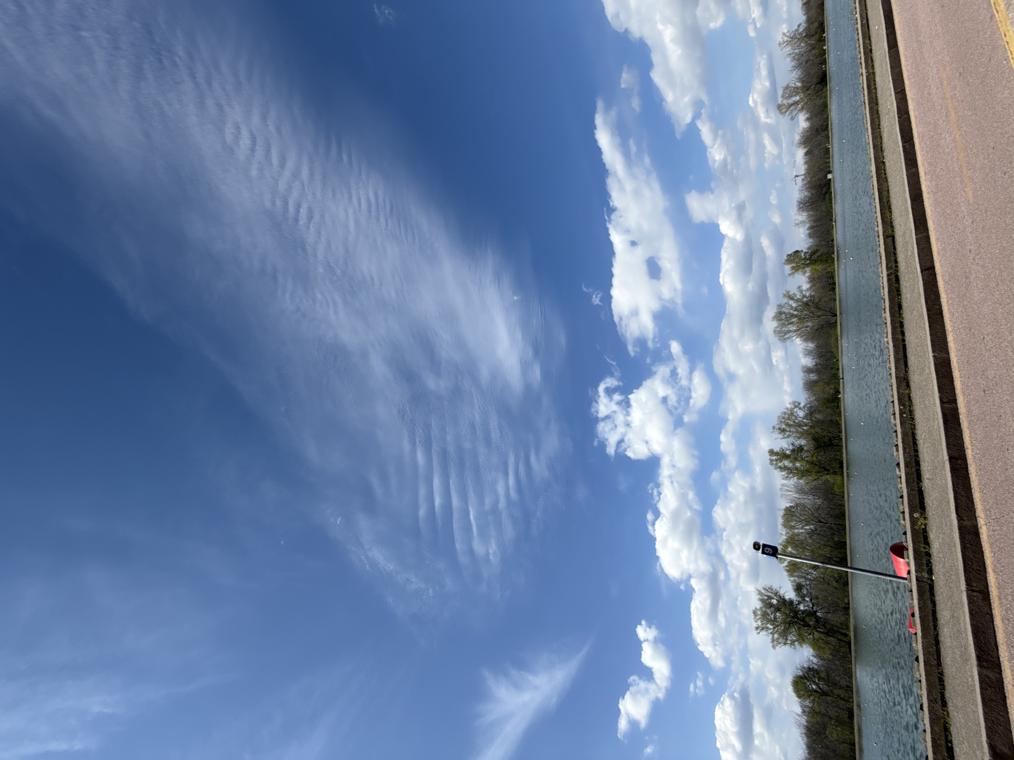 The image depicts a canal scene with a blue sky containing cirrus and cumulus clouds. The canal is bordered by a concrete and asphalt path. Trees line the opposite bank of the canal. A metal pole with two black rectangular signs is situated near the water's edge. A red object is visible next to the pole. The image appears to be taken on the side of the canal on the pathway. The precise location within Plovdiv is not discernible from the image alone.
