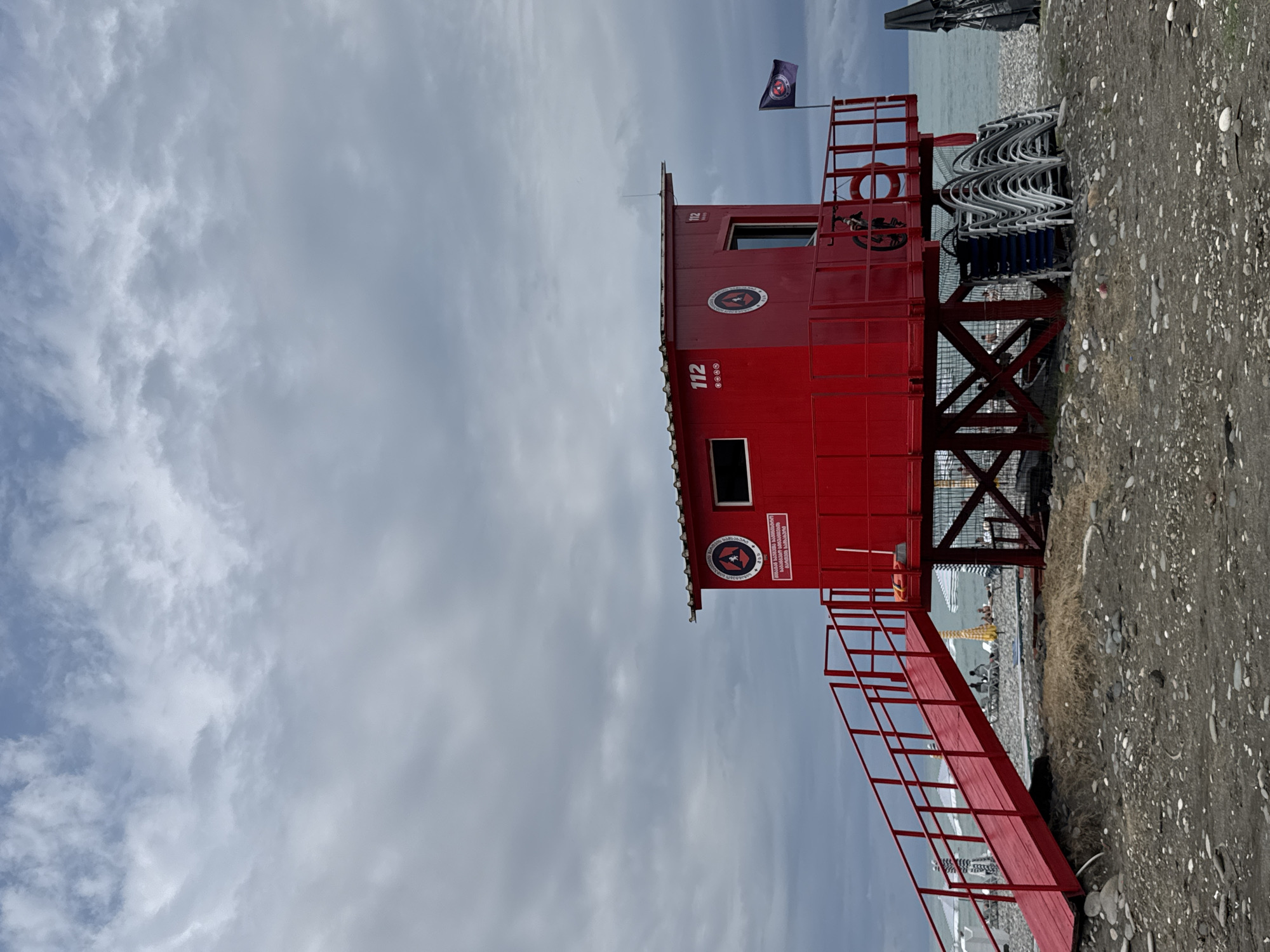 The image shows a bright red lifeguard tower on a pebble beach in Batumi, Georgia. The tower has a ramp leading up to it, a small window, and a door. There are several logos and numbers visible on the tower, including the number "112" and a circular emblem. A dark flag with a logo is flying from a pole next to the tower. In the background, the sea stretches out under a cloudy sky. There are some beach chairs stacked near the base of the tower, and a few people can be seen in the distance on the beach. The overall scene suggests a typical beach day, with the lifeguard tower serving as a prominent feature of the coastal landscape.